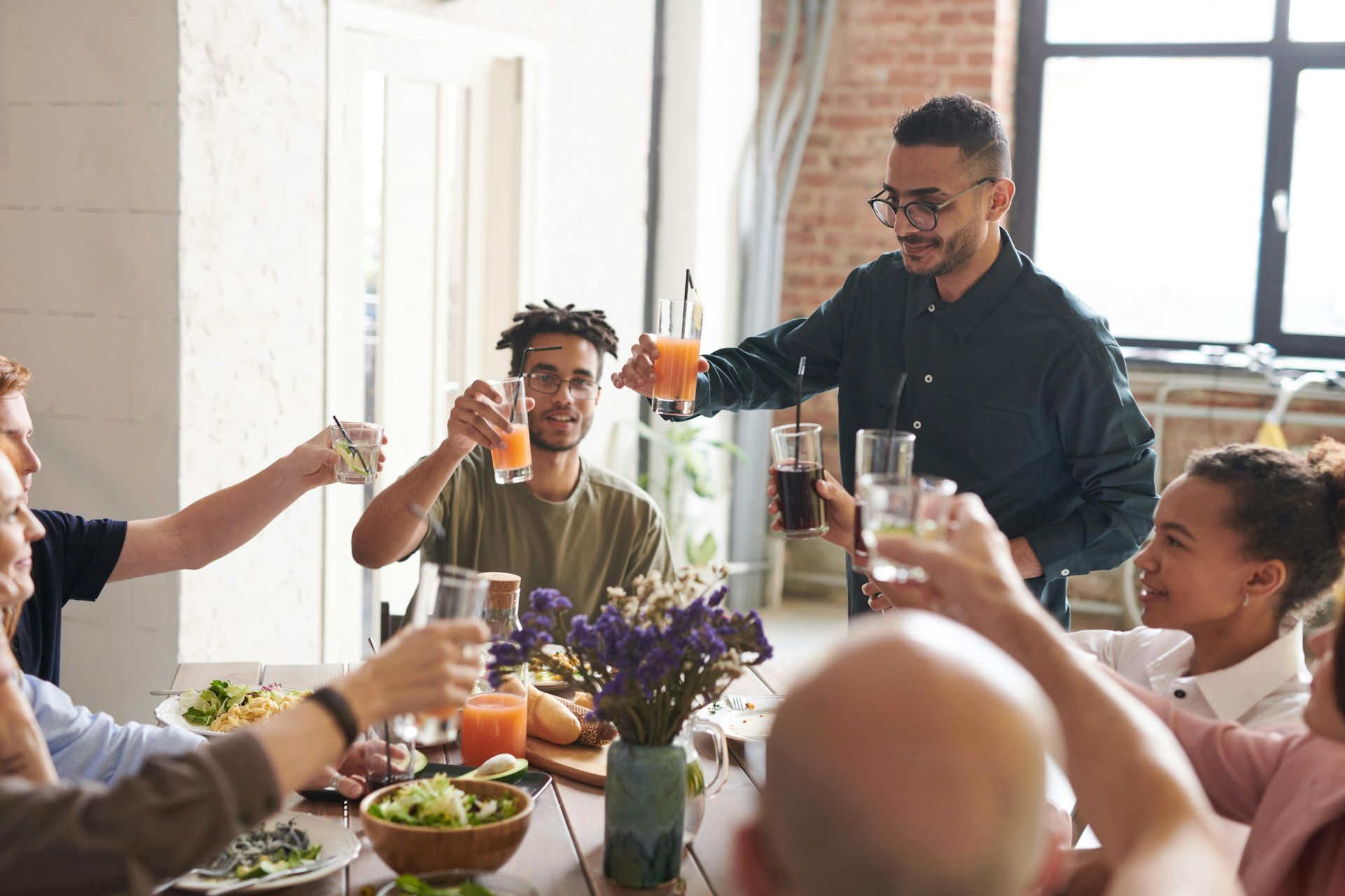 People toasting drinks around a table at a gathering; food and flowers present.