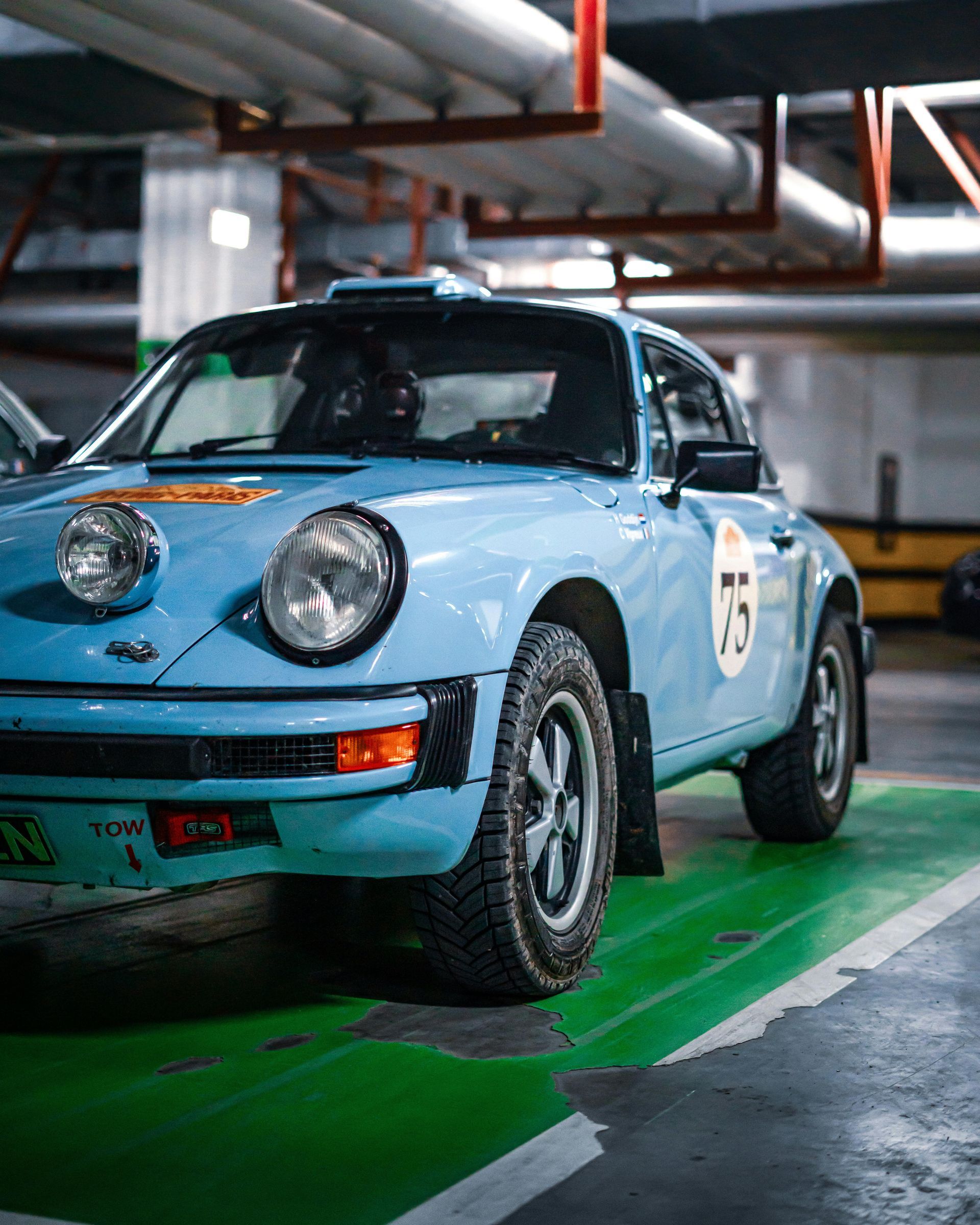 Light blue Porsche rally car with number 75 parked on a green surface in a parking garage.