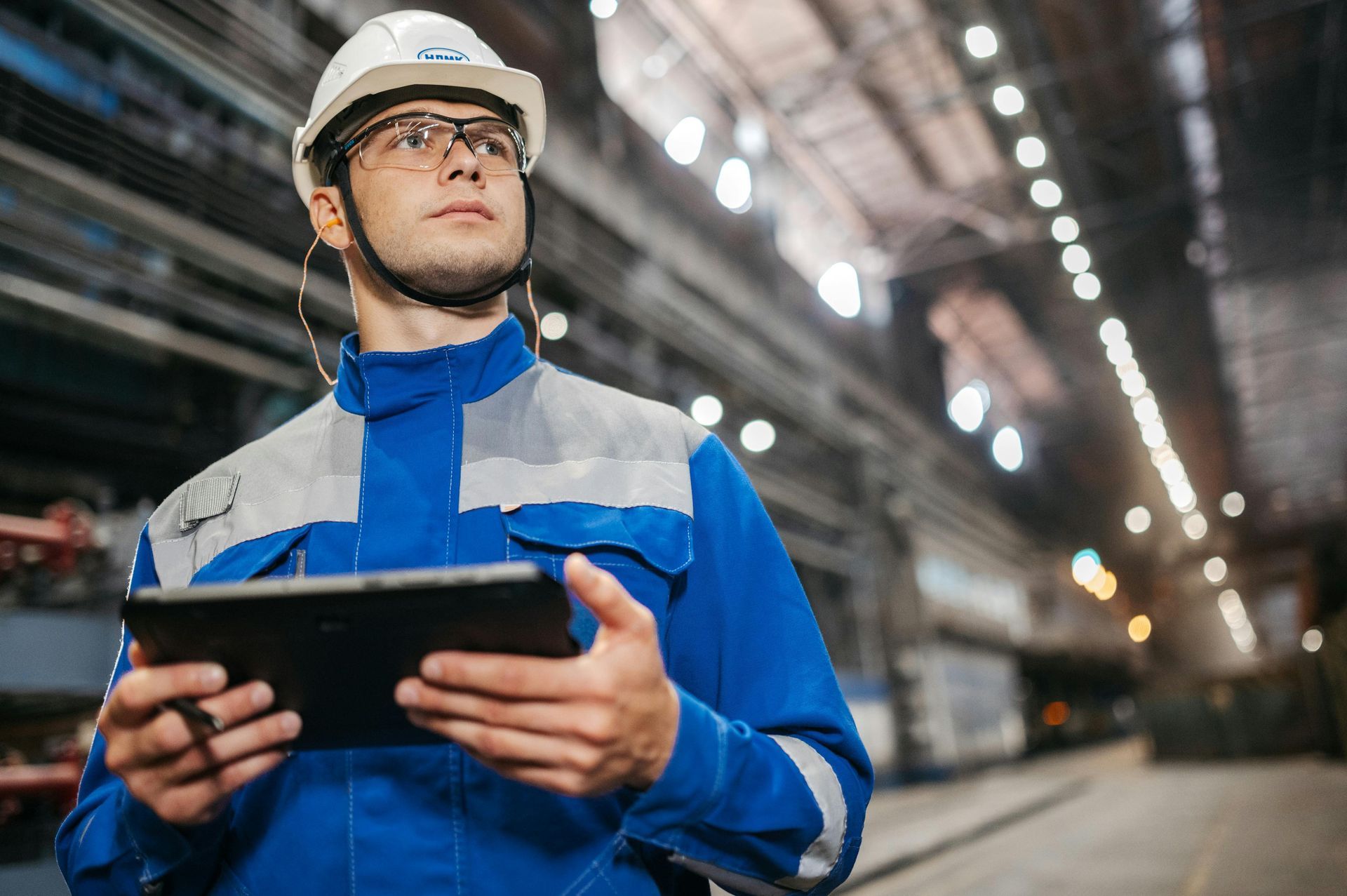 Man in blue workwear and hardhat holding a tablet, looking up in a factory setting.
