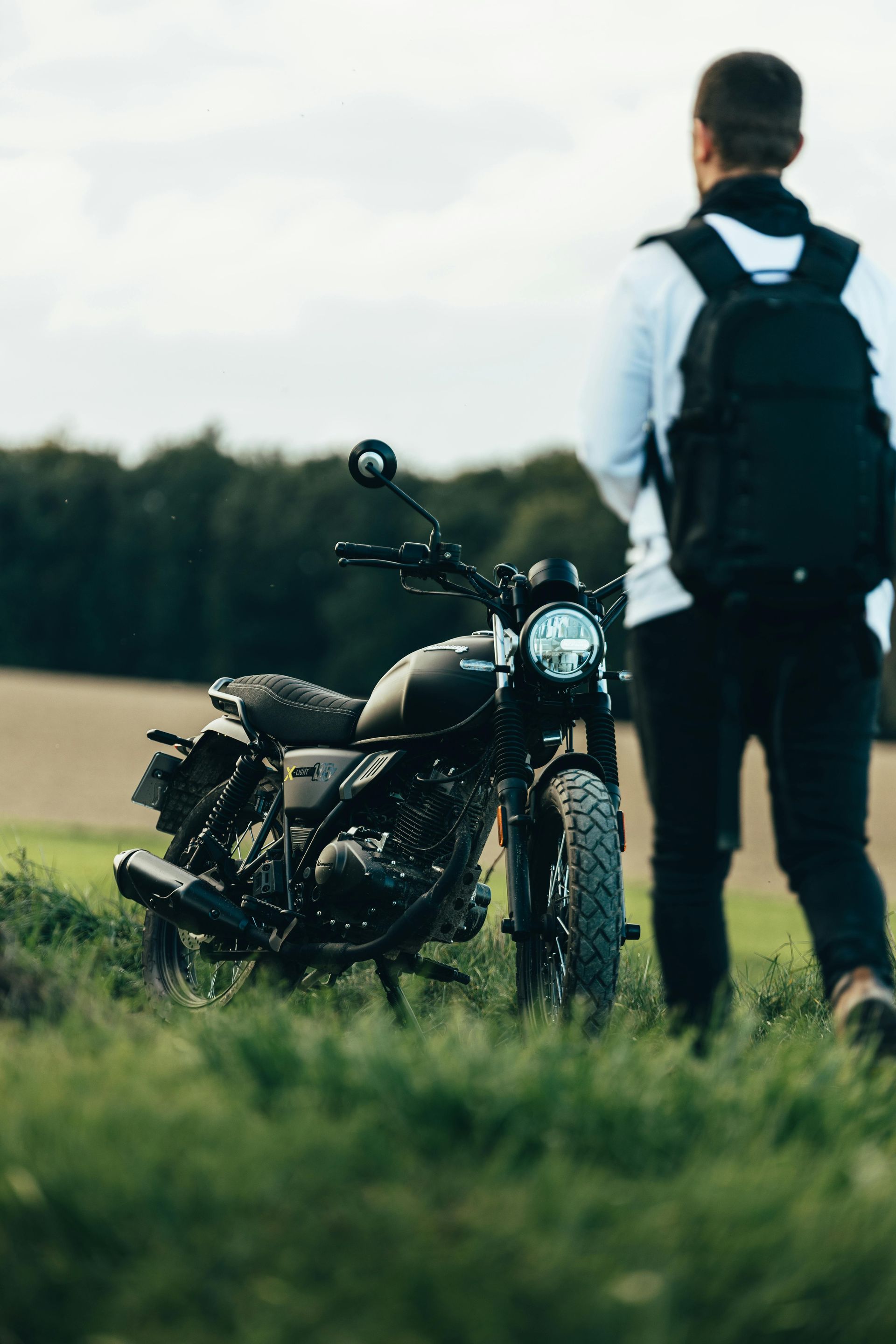 Black motorcycle parked in grassy field; person with backpack walks away.