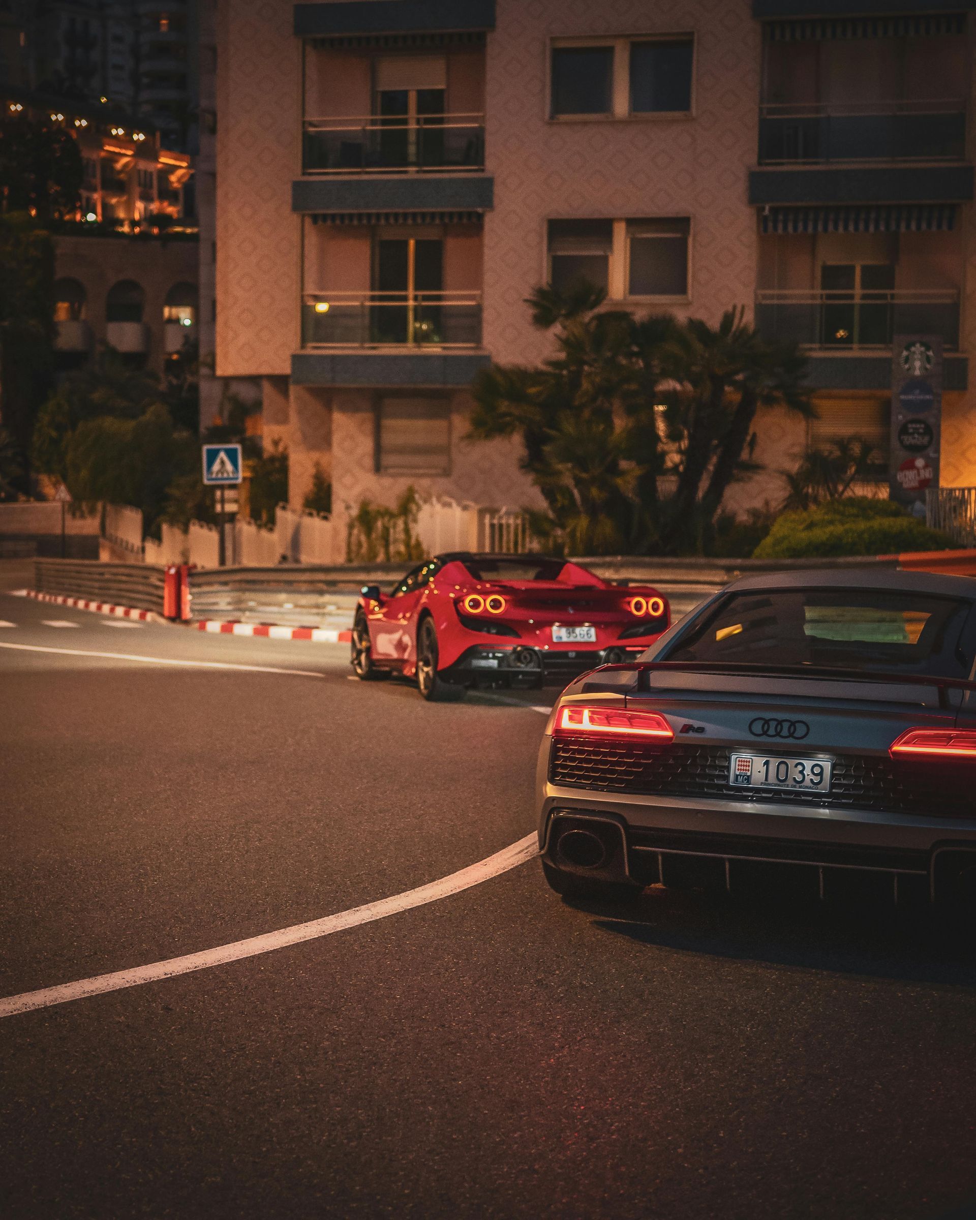 Two sports cars, a red Ferrari and a grey Audi, on a city street at night.