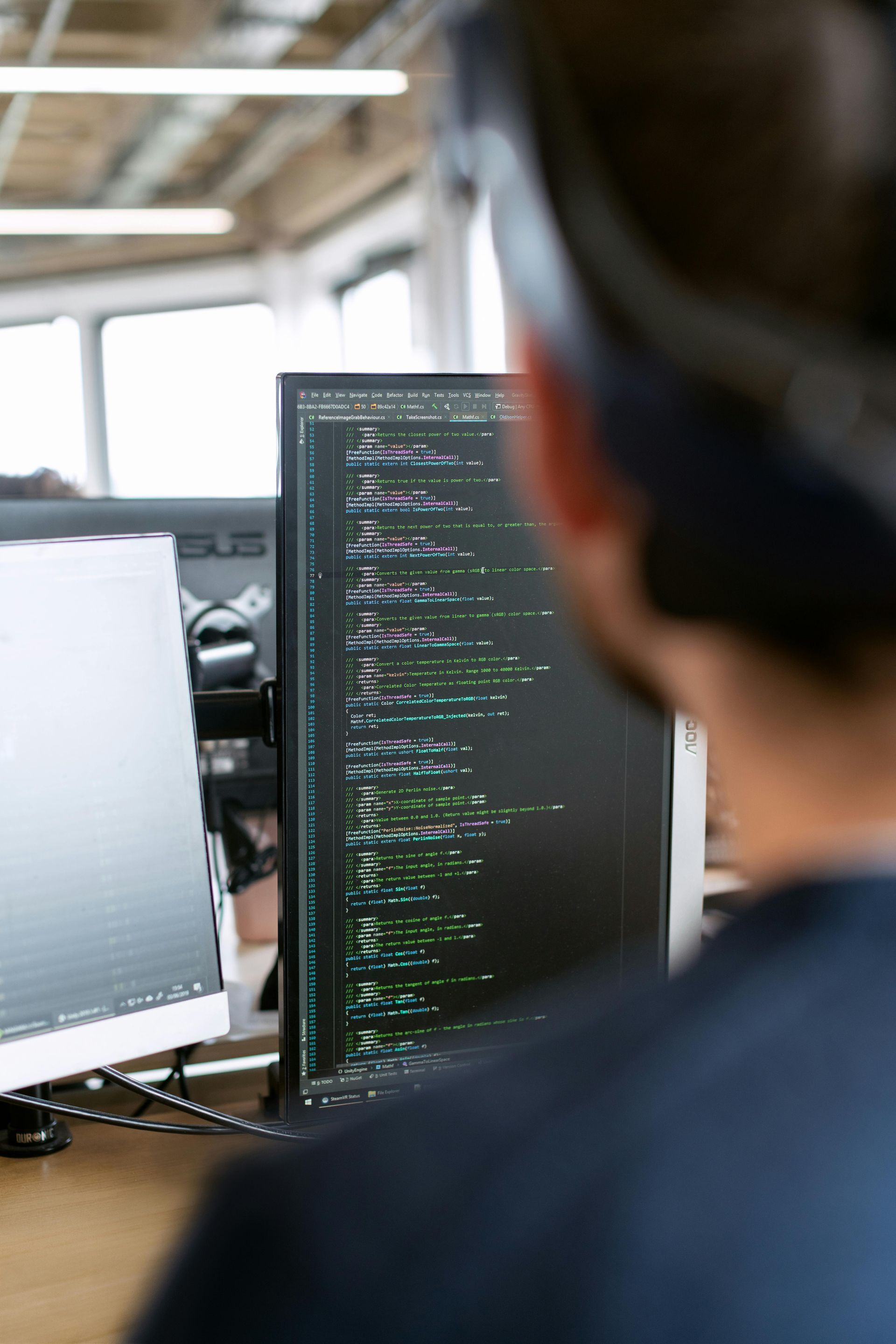 Person coding on a computer, with code displayed on the monitor. Another monitor is visible.