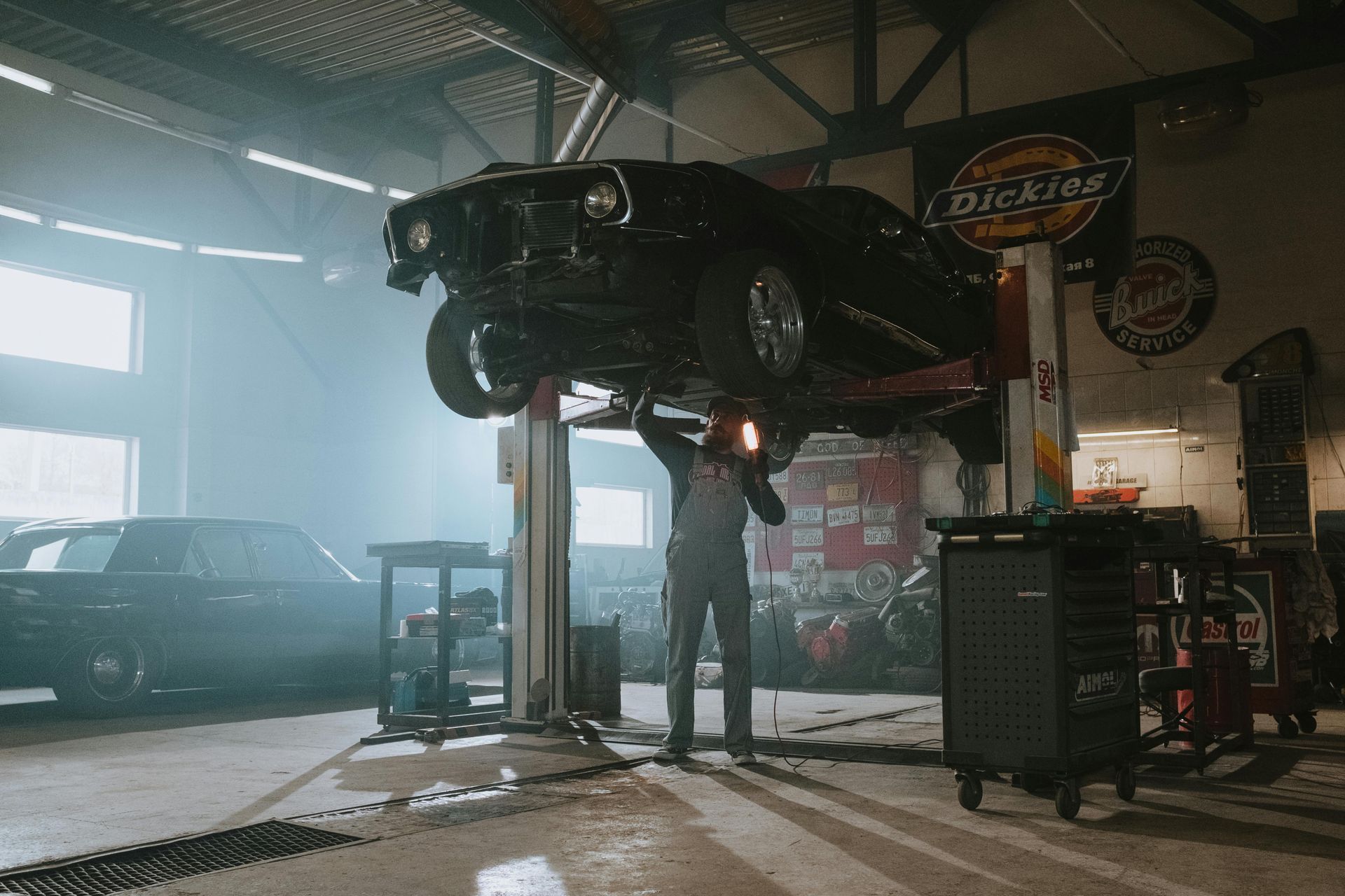 Mechanic working on a black car lifted in a garage. Dusty environment, Dickies sign.