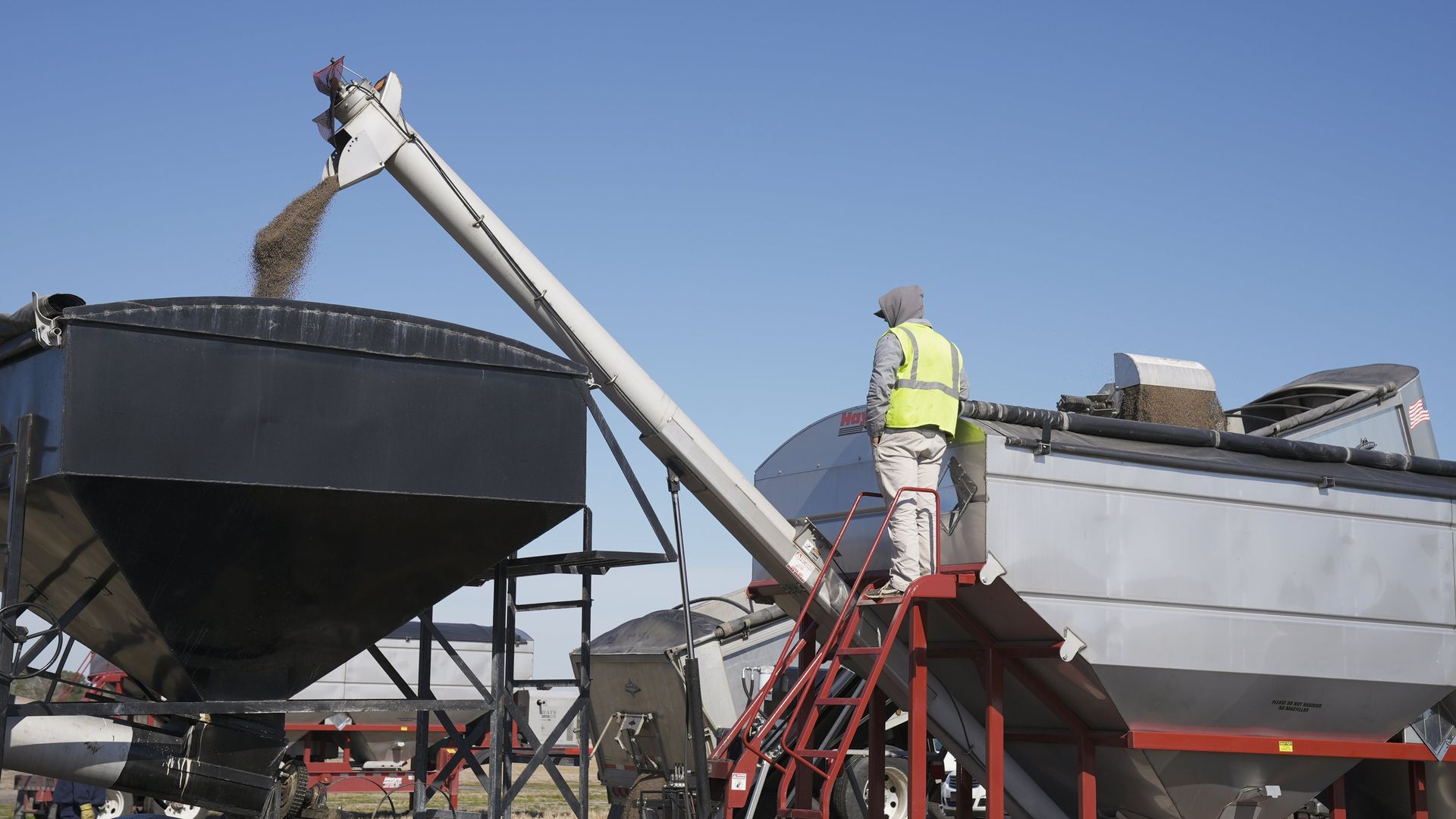 A worker in a yellow vest oversees grain being unloaded from an auger into a silo outdoors.