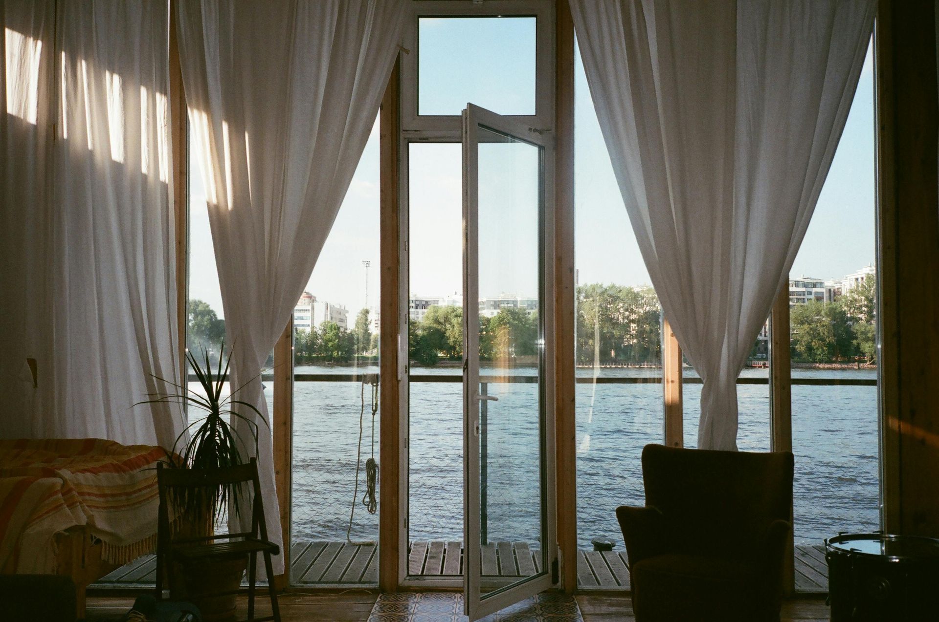 Interior view of a room with large windows overlooking a river, white curtains, and minimal furniture.