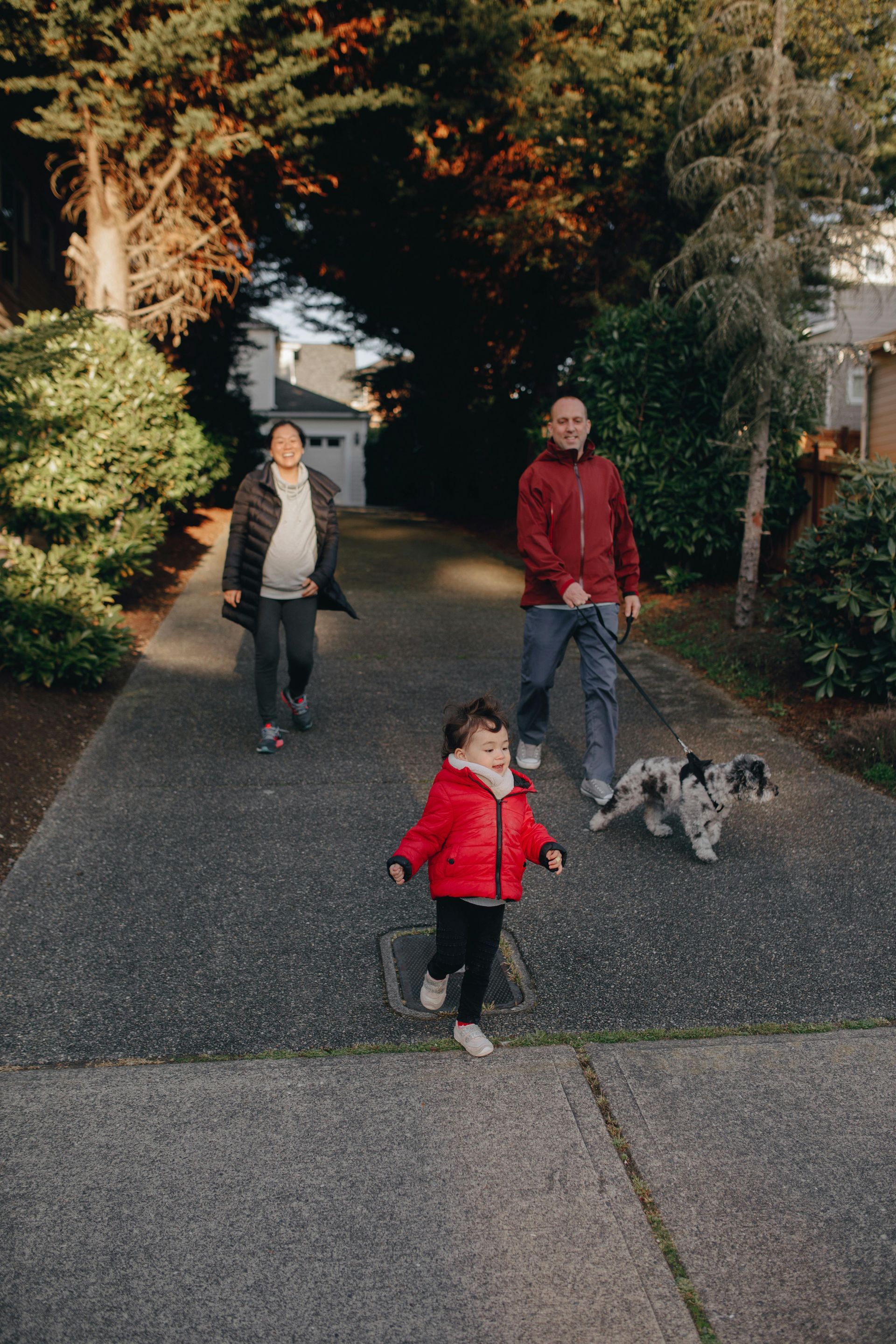 Family walks on a paved path: Child runs, parent walks dog, parent follows. Green trees and houses surround.