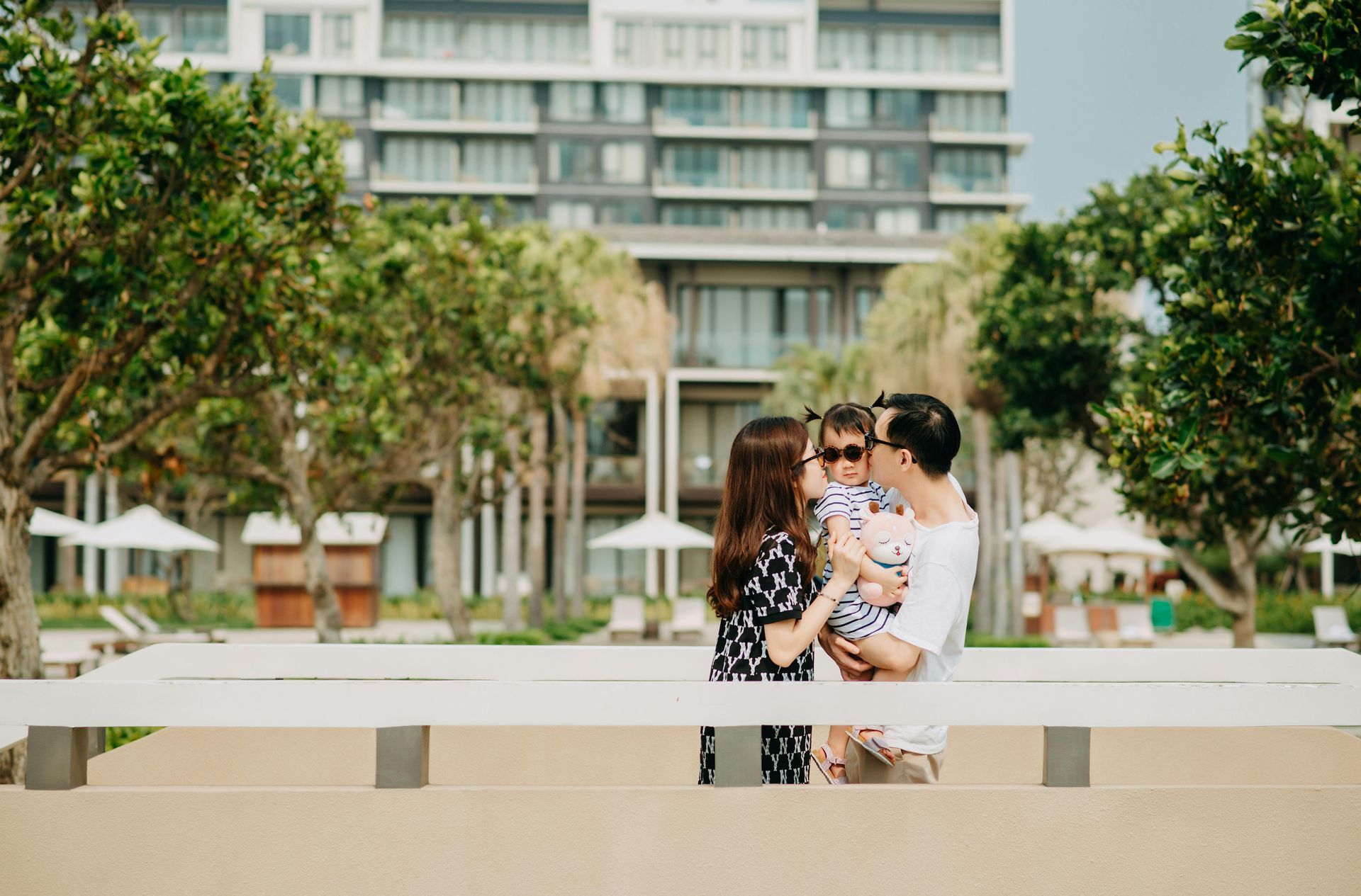 Family kisses in front of resort buildings. Woman in black dress, man in white shirt holding child.