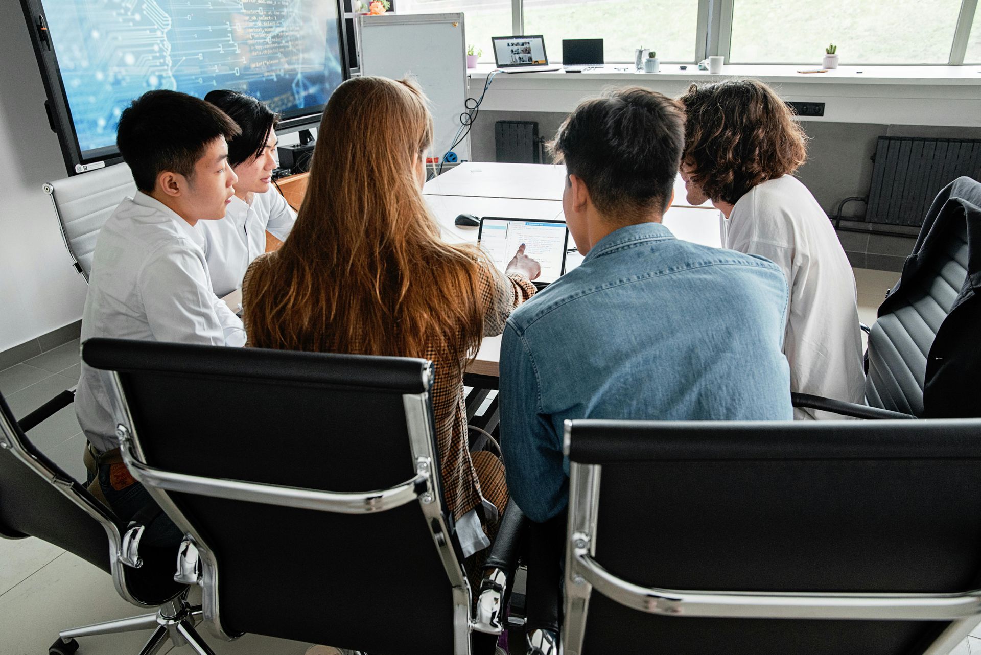 Five people in a meeting, gathered around a table, with a large screen in the background.