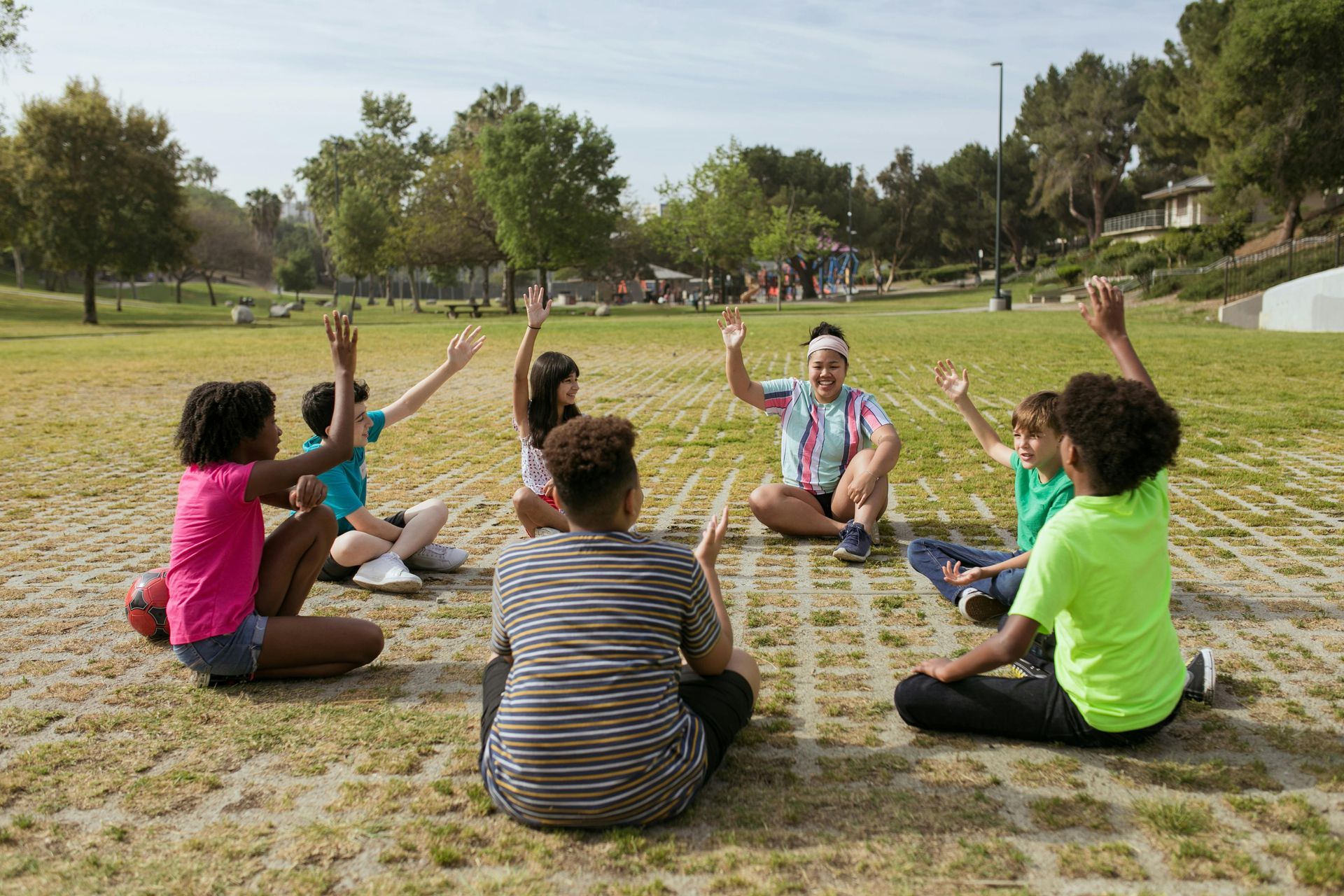 Children and an adult sit in a circle on a grassy field with hands raised. Sunny day.