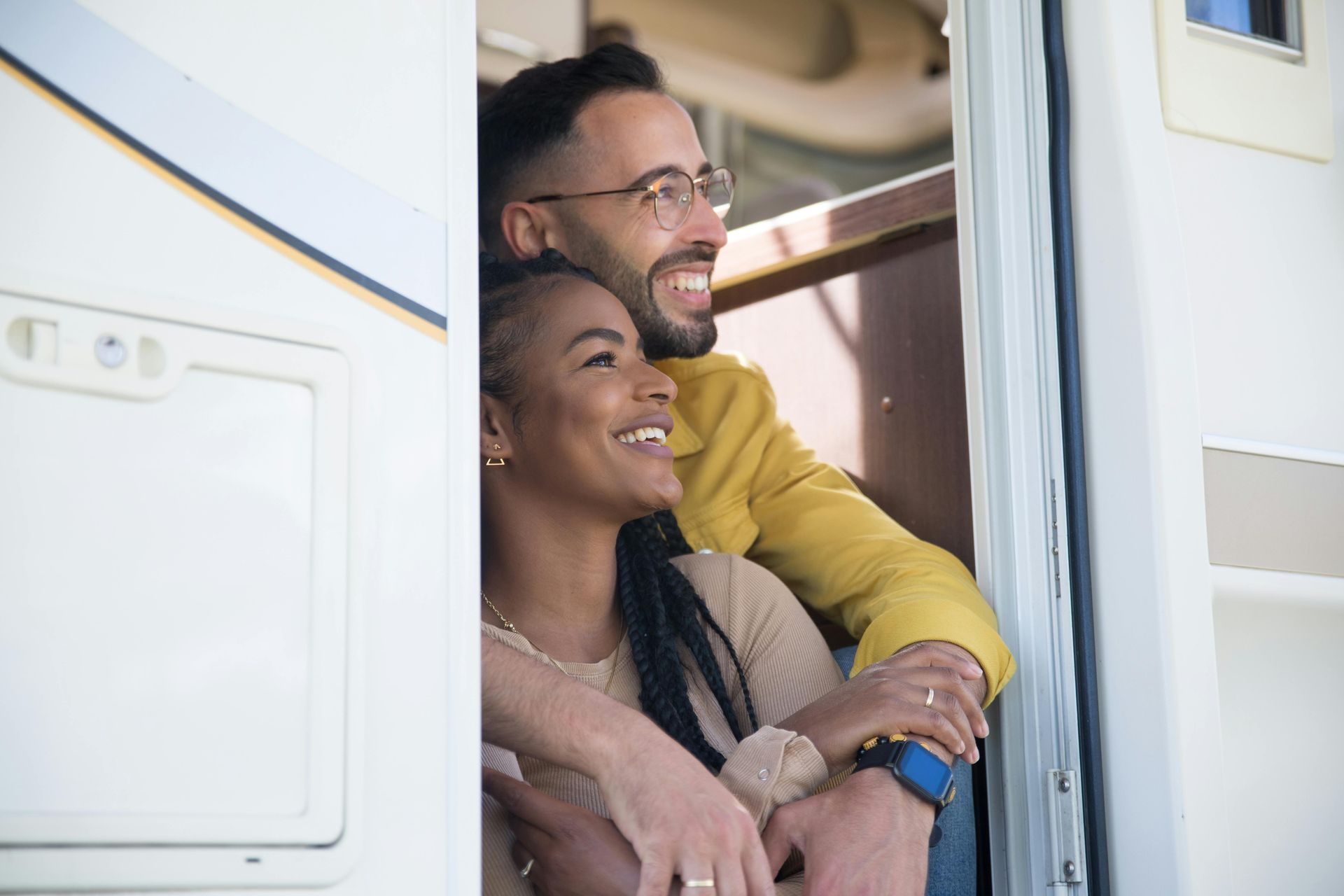 Couple embraces looking out from a recreational vehicle. Smiling, man wears glasses and yellow jacket. Woman has long braids.