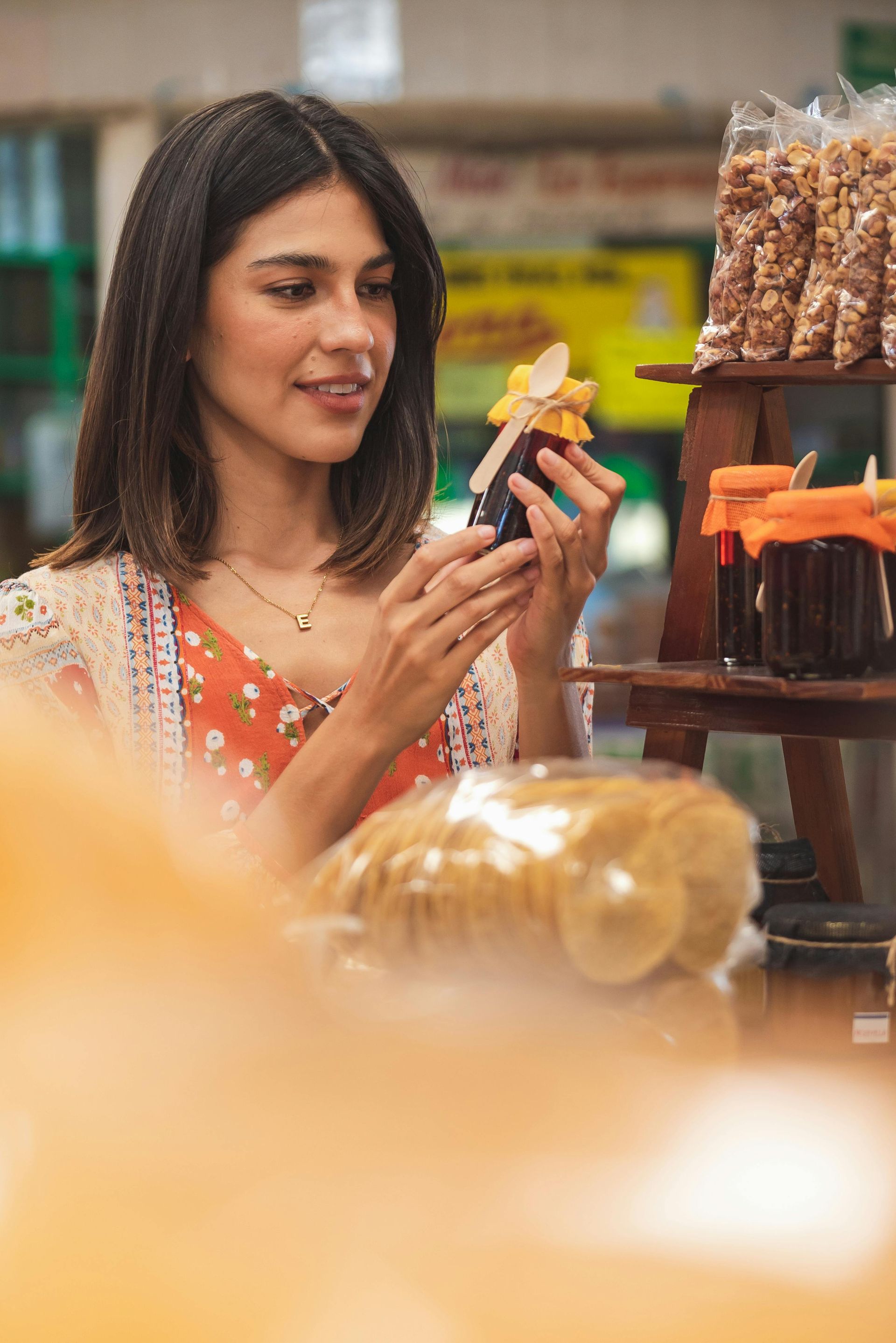 Woman examines jar of preserves at a market, smiling, wearing a floral dress and shawl.