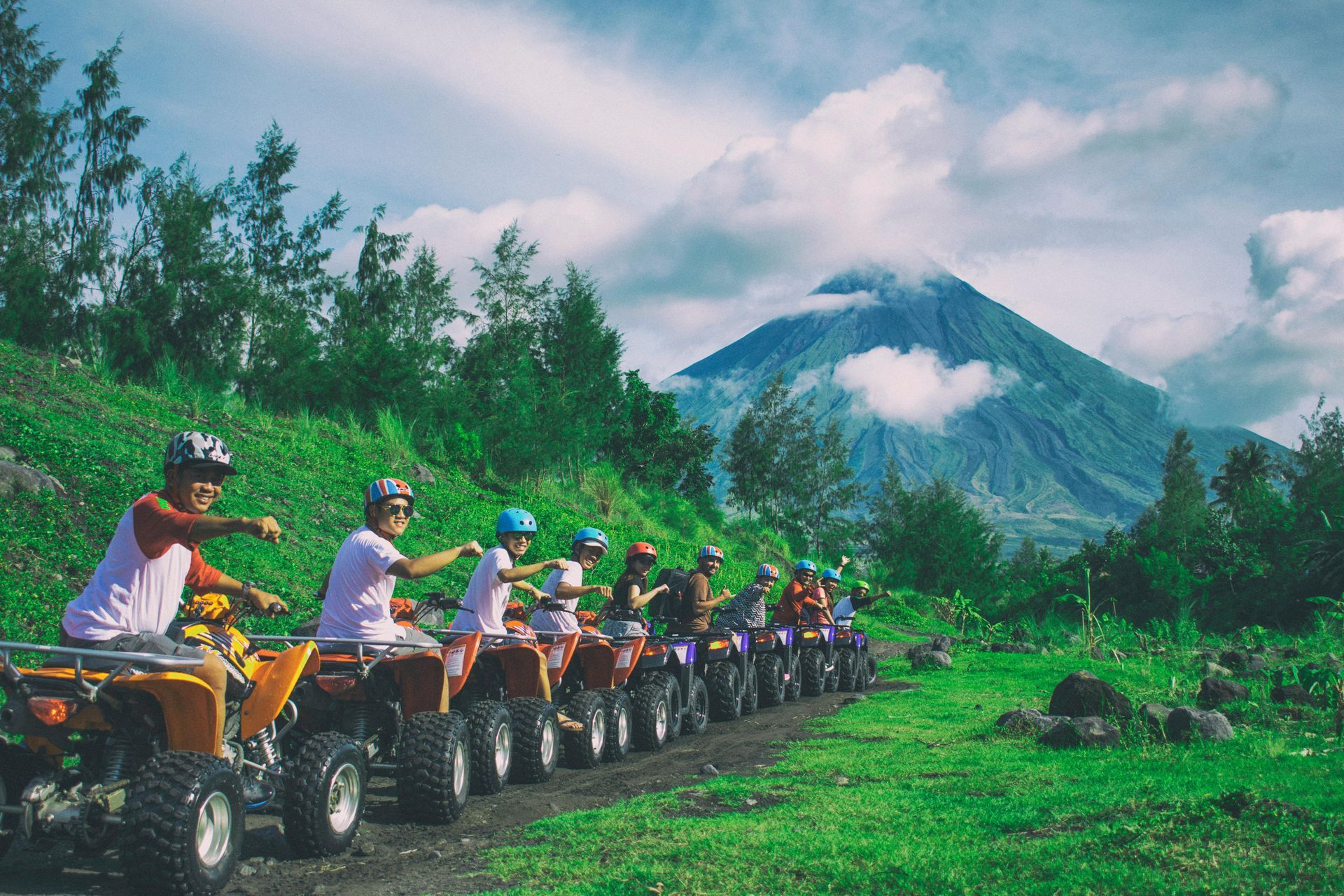 People riding ATVs on a green path with a mountain in the background, cloudy sky.