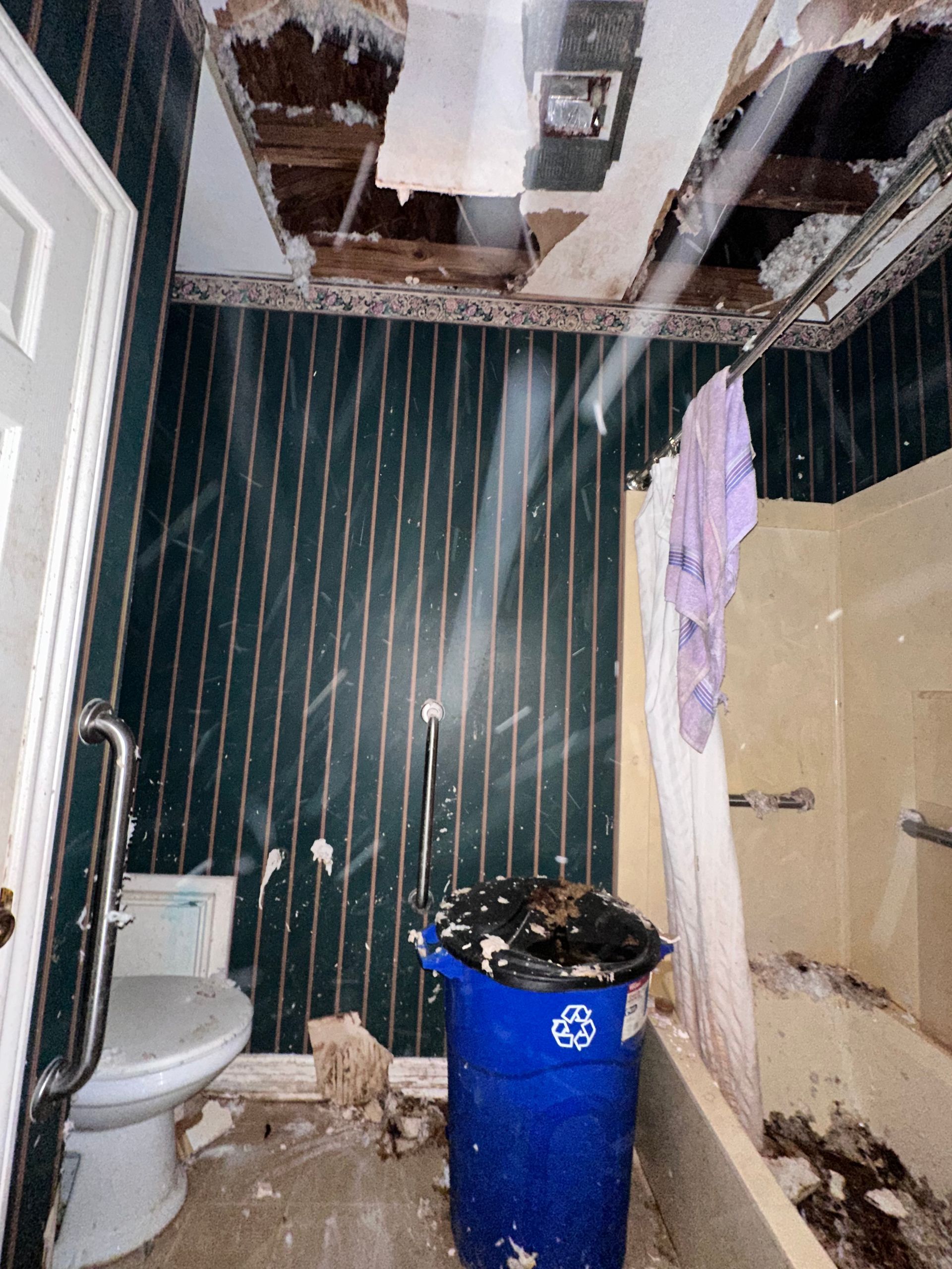 Damaged bathroom interior with exposed ceiling, peeling wallpaper, and blue trash can.