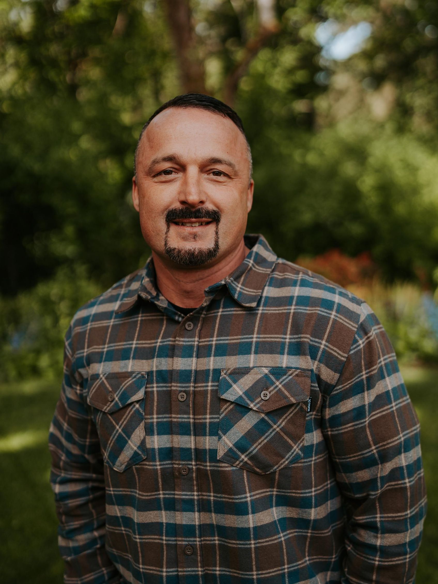 A man with a beard is wearing a plaid shirt and smiling.