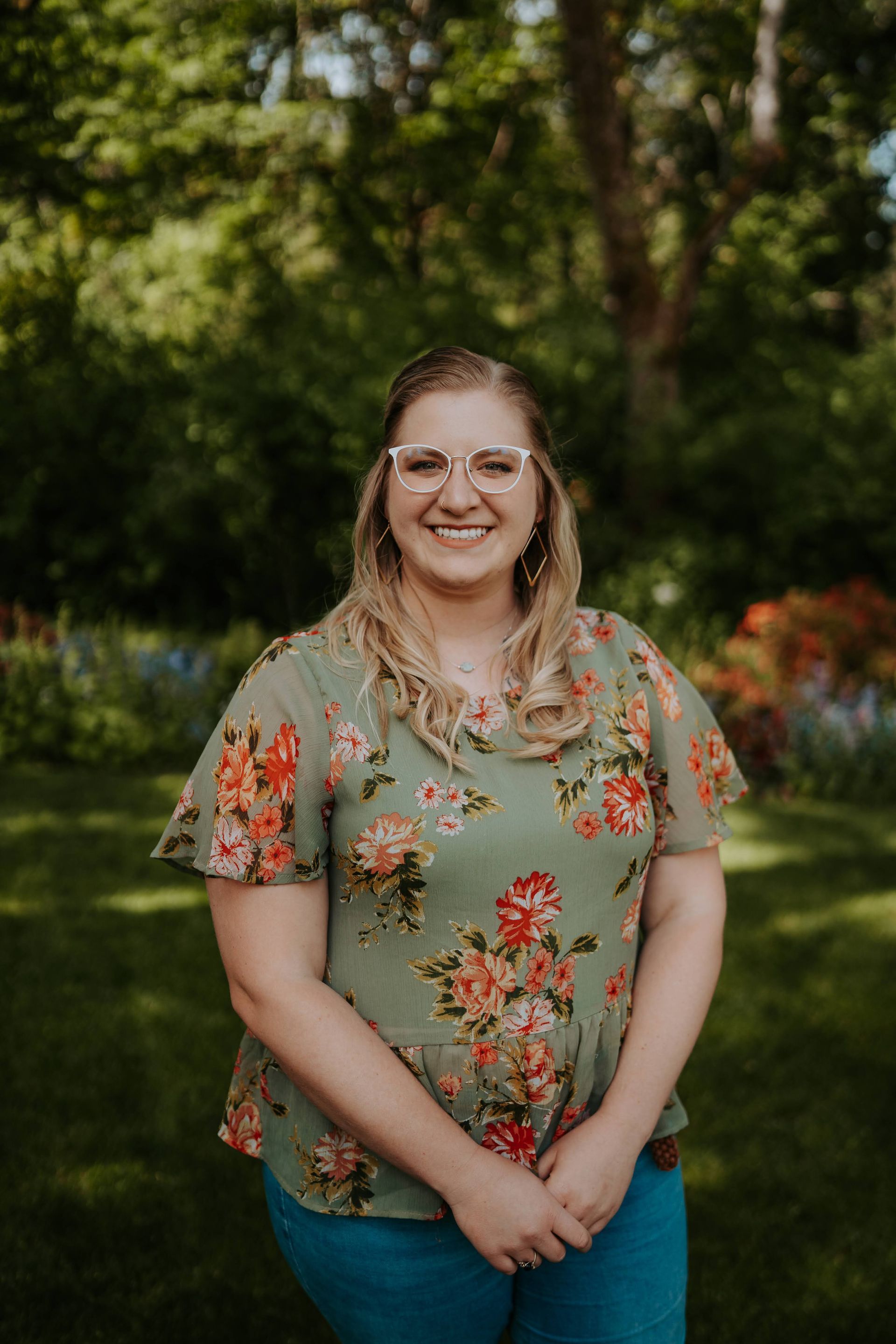 A woman wearing glasses and a floral shirt is standing in a field.