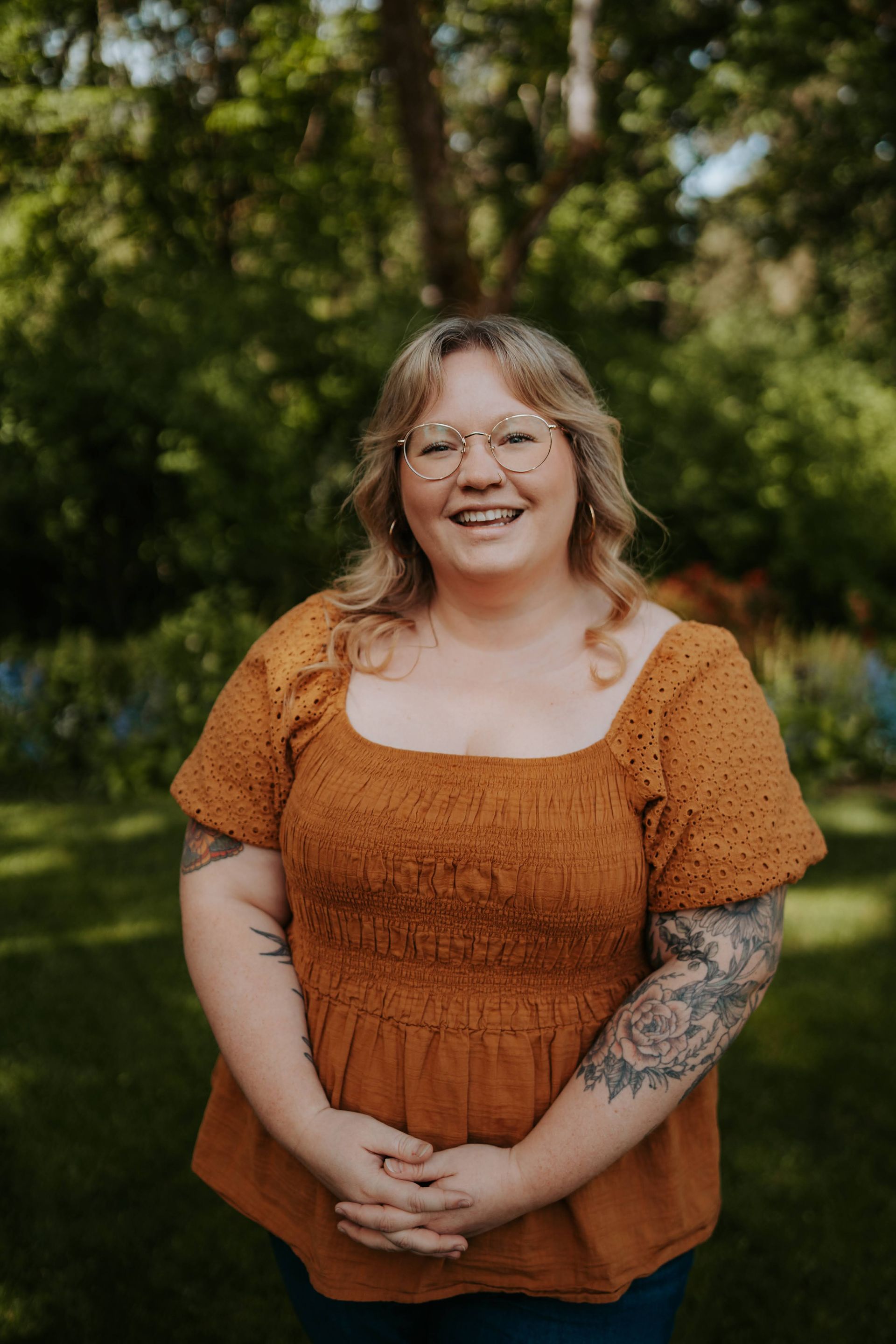 A woman wearing glasses and a brown top is standing in a park with her hands folded and smiling.