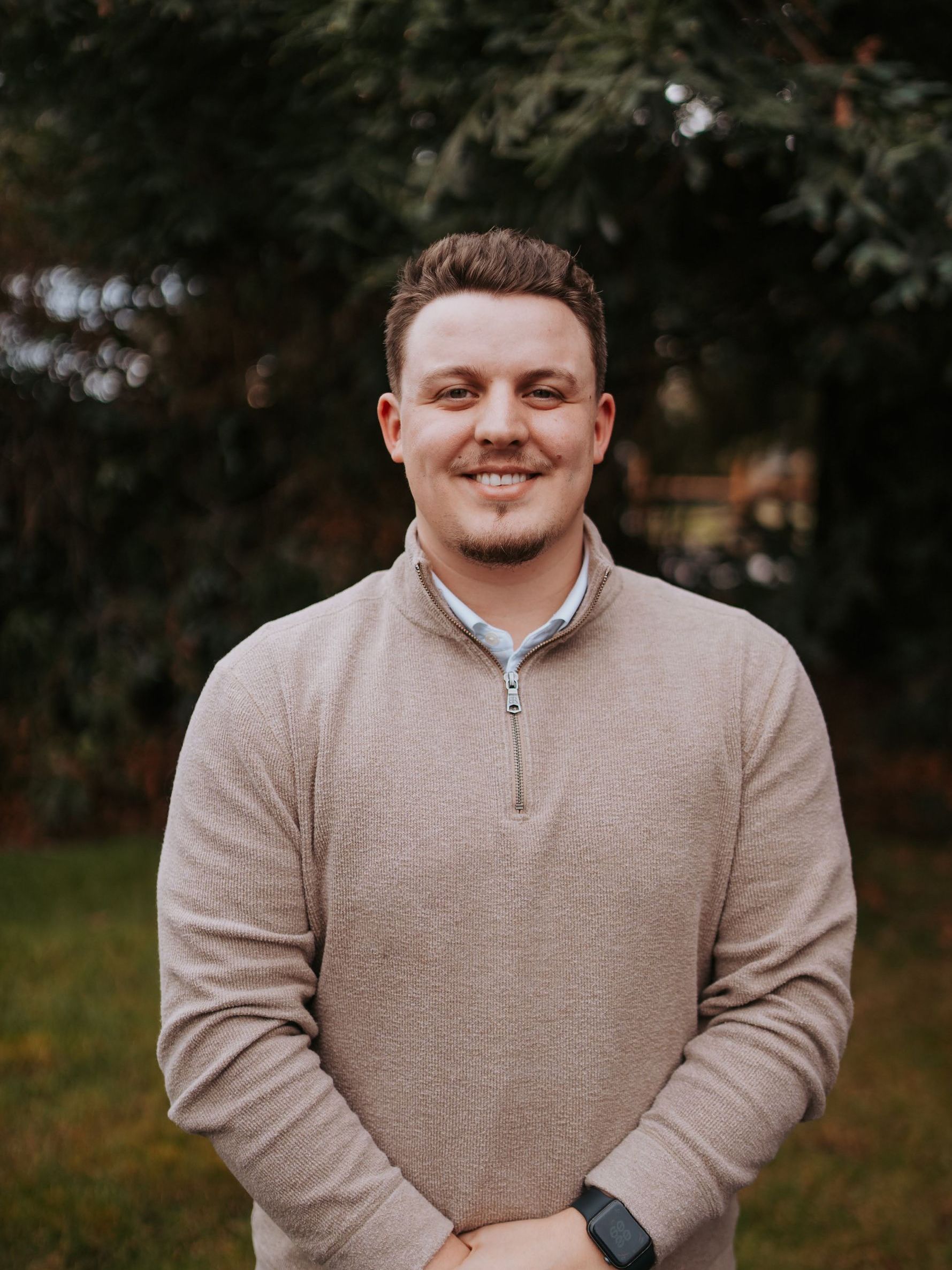 A man in a tan sweater is smiling for the camera while standing in front of trees.