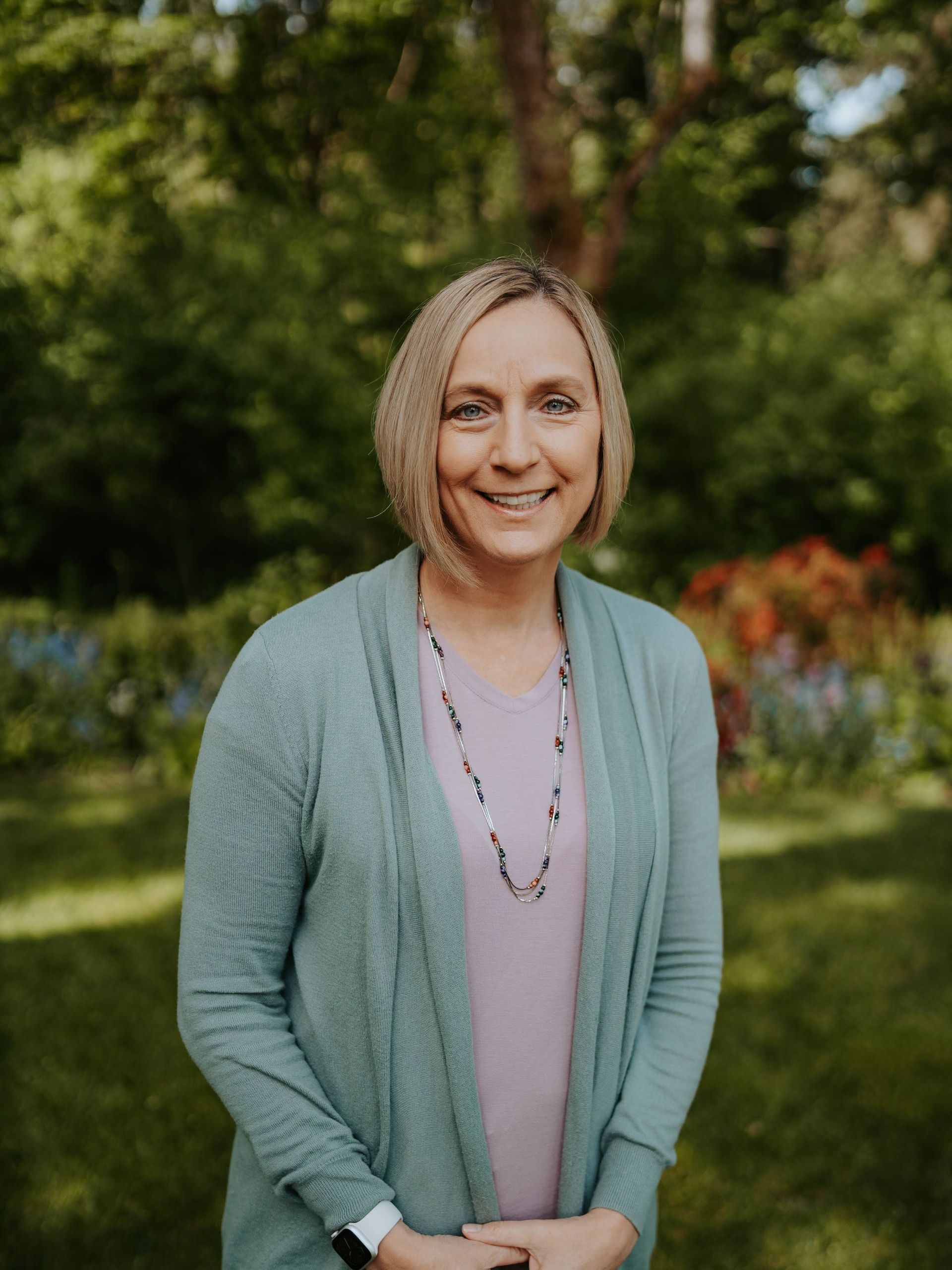 A woman wearing a green cardigan and a necklace is standing in a park.
