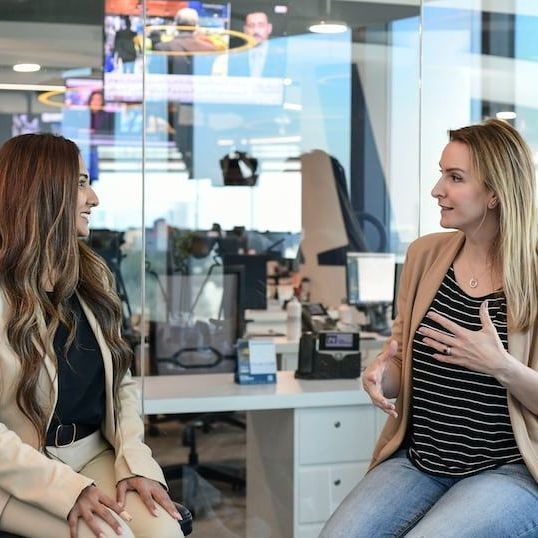 Two women are sitting in front of a glass wall talking to each other.