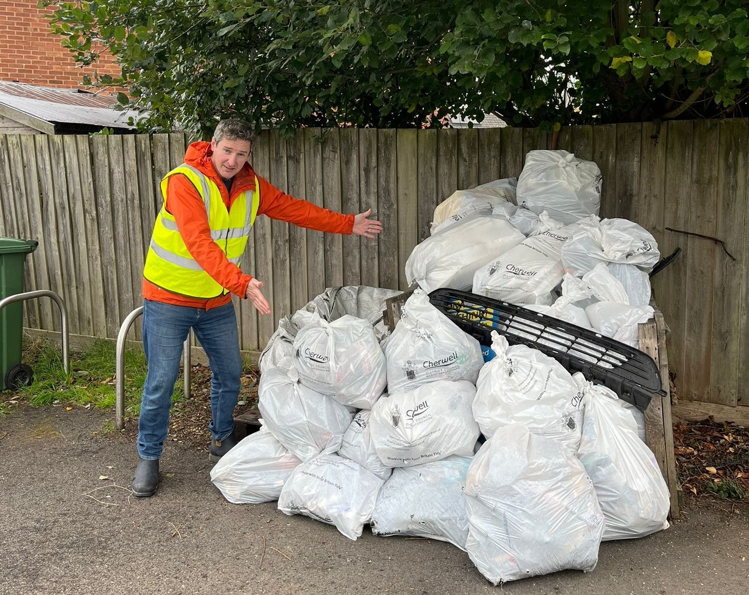 Councillor Samuel Smith in vest points at pile of white trash bags in front of a wooden fence and green tree.