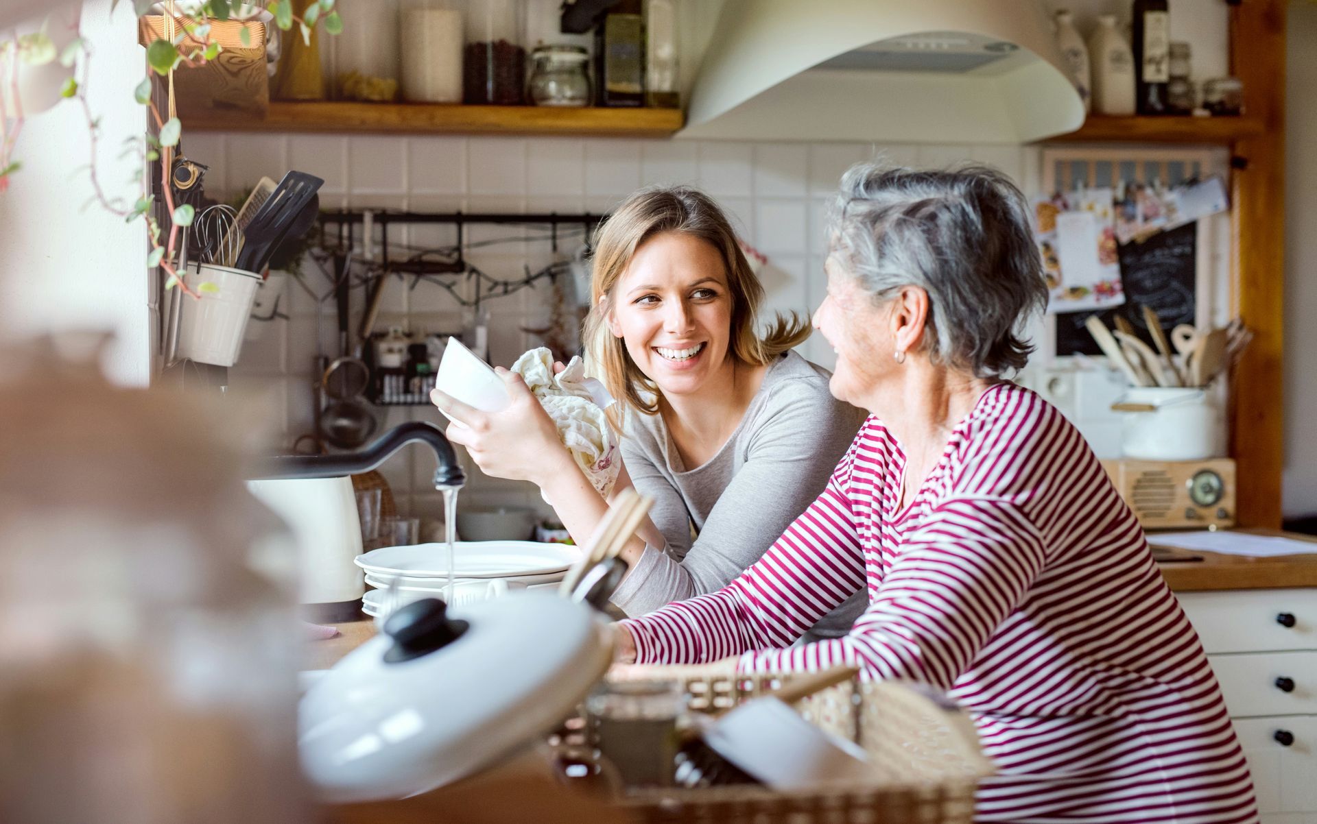 Woman washing dishes with older woman in a kitchen; smiling, wooden shelves, white cabinets.