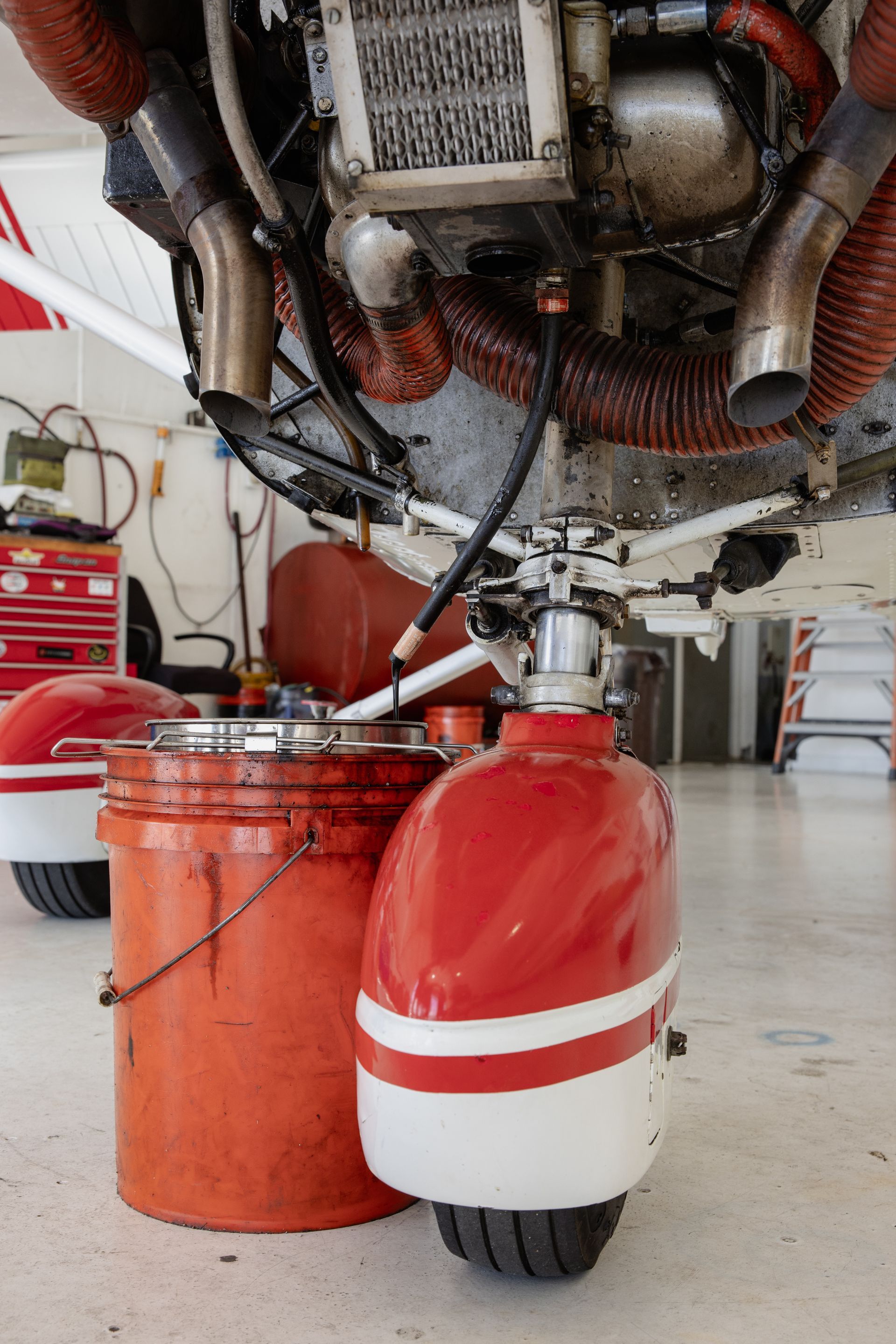 Two fuel containers beneath an aircraft engine, one red-white and one orange, in a hangar.