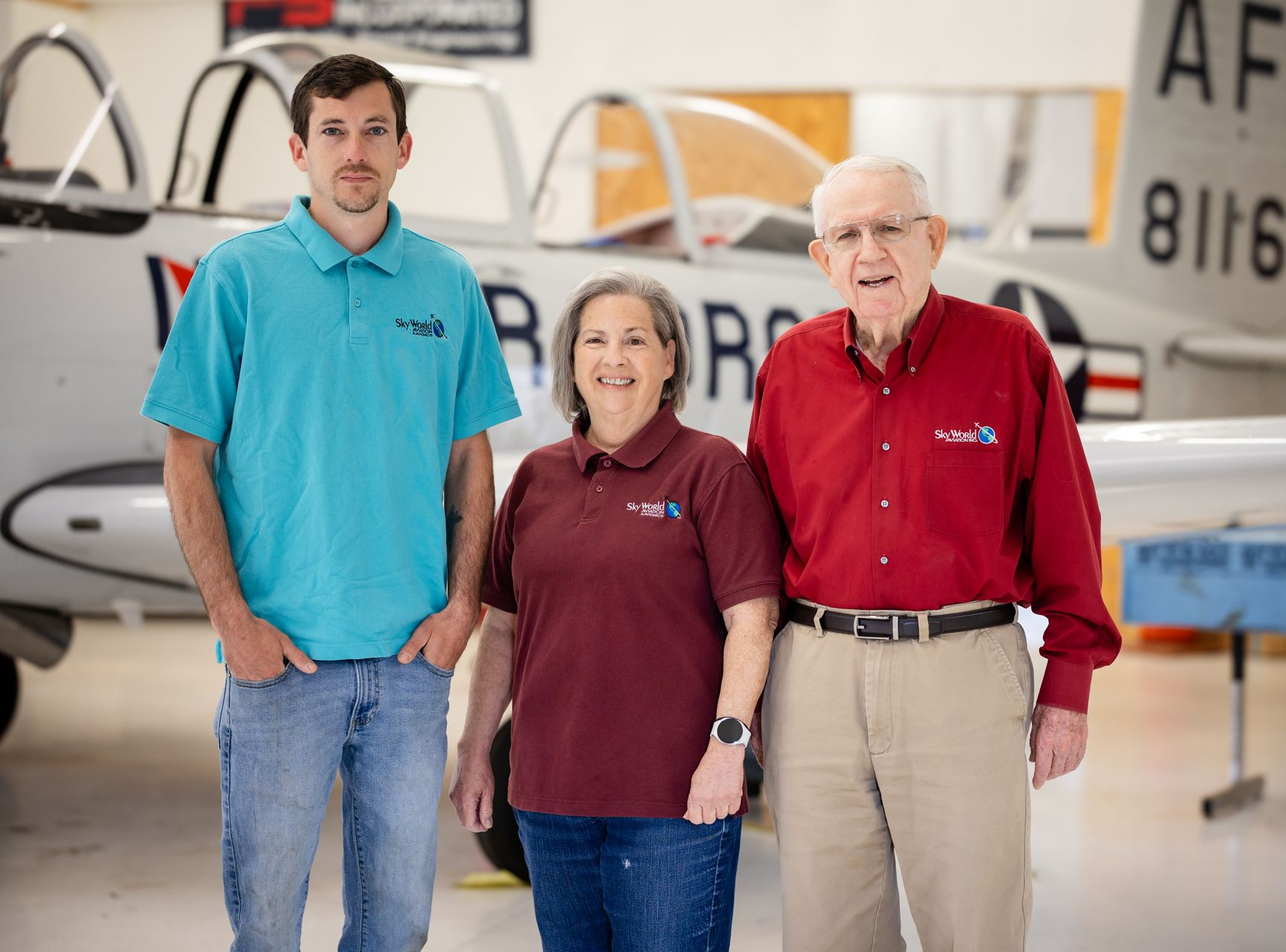 Three people standing in a hangar with small airplanes, wearing teal, maroon, and red shirts.