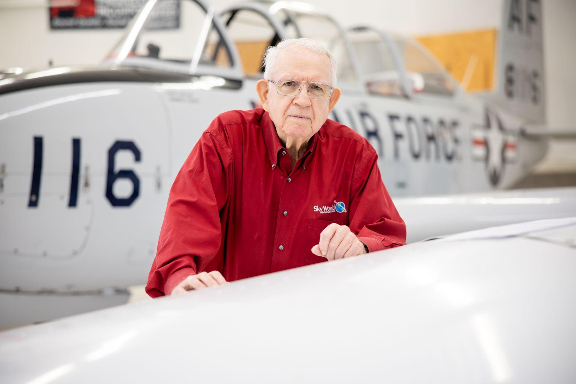 Man in red shirt leaning on a white airplane in an aviation hangar