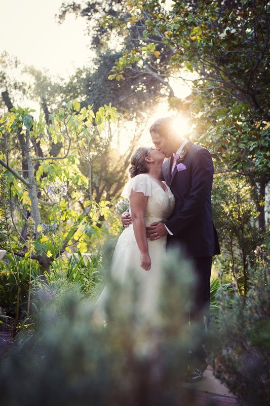 A bride and groom are kissing in a garden with the sun shining through the trees.