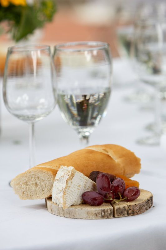 A wooden cutting board with bread , cheese , grapes and wine glasses on a table.