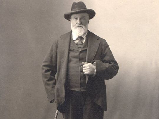A man with a beard is wearing a hat and holding a cane.