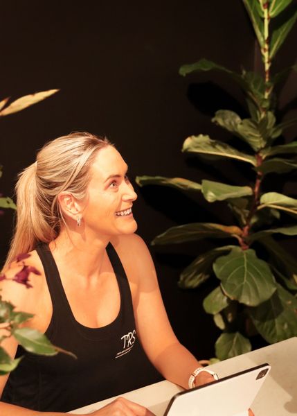 Image depicts Head TRS Pilates Instructor Ella welcoming a client at the TRS Pilates Ballarat Studio. She is sitting at a marble desk with an Ipad, wearing a TRS top with a large fig plant in the background.