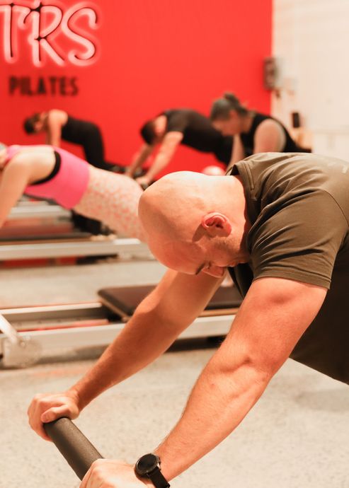 Image is taken inside the TRS Pilates Ballarat studio. It depicts 5 people performing Reformer Pilates exercises in the form of a plank. There is a red wall with letters TRS fixed to the wall with a lighting background and the word Pilates in black fixed below. There are several Reformer Pilates machines in the studio being used by clients and the vibrant polished concrete floor is on display.