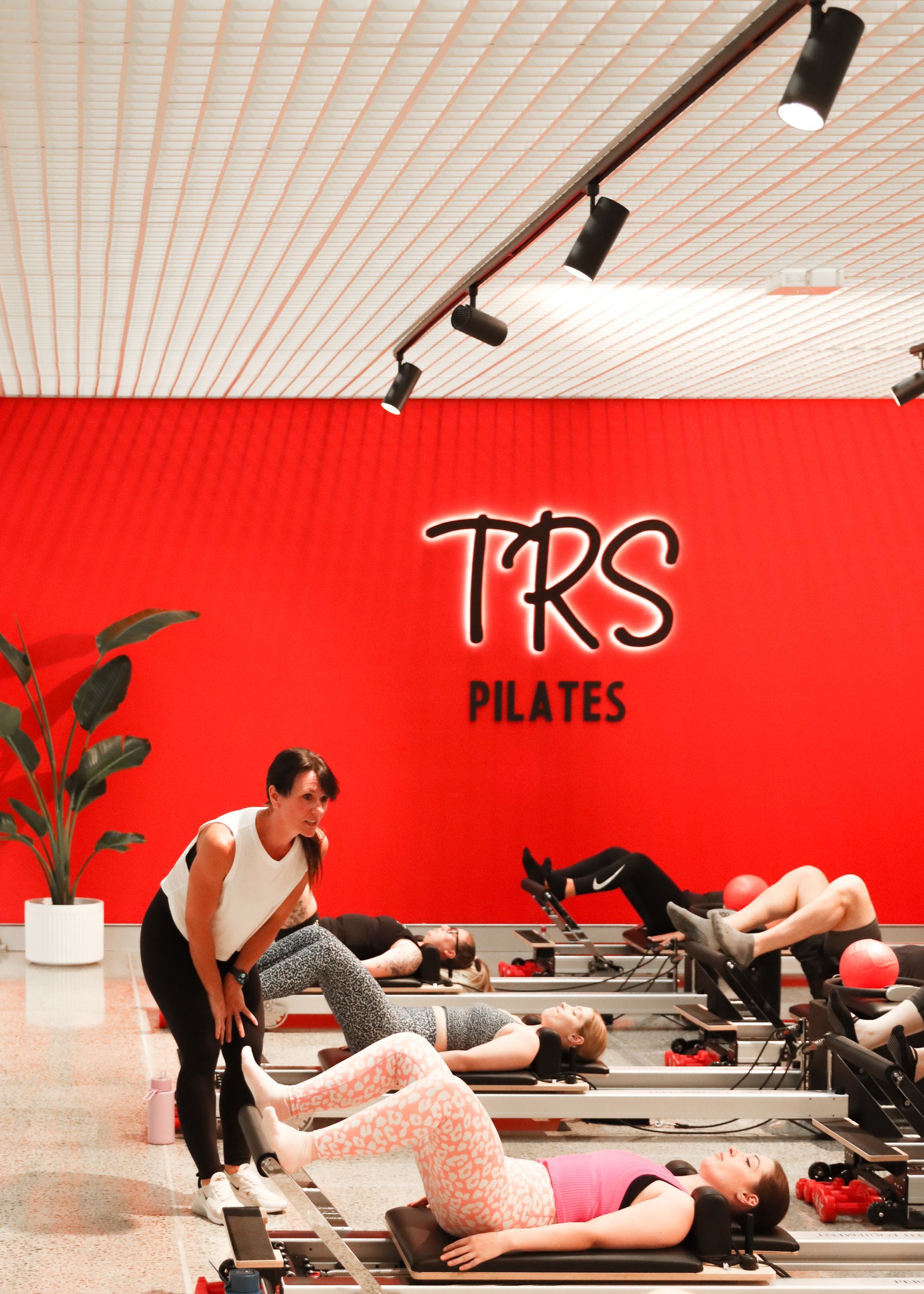 Image depicts the TRS Pilates Ballarat studio featuring the spectacular down lights, red feature wall with TRS lighted signage, large green fig plant in the corner, with at least 6 clients participating in a Reformer Pilates Class, whilst a senior instructor teaches the class.