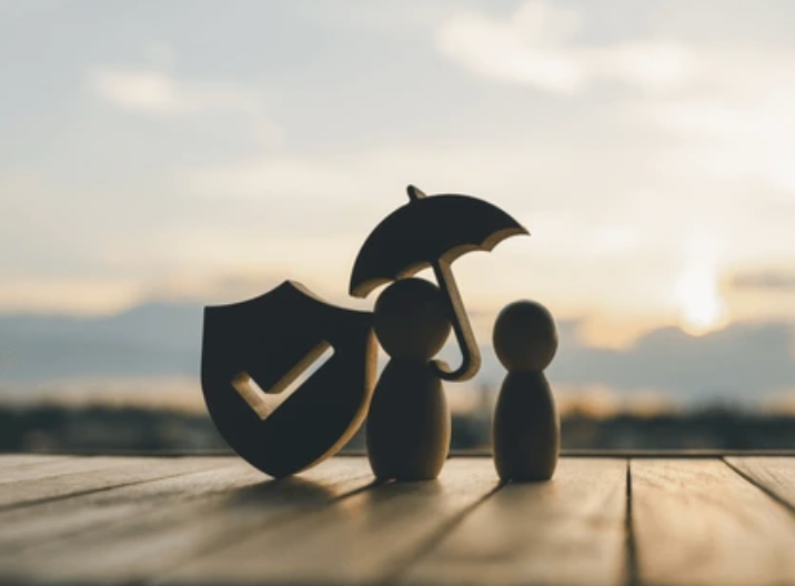 Wooden figurines under an umbrella and a shield with a checkmark, symbolizing protection.