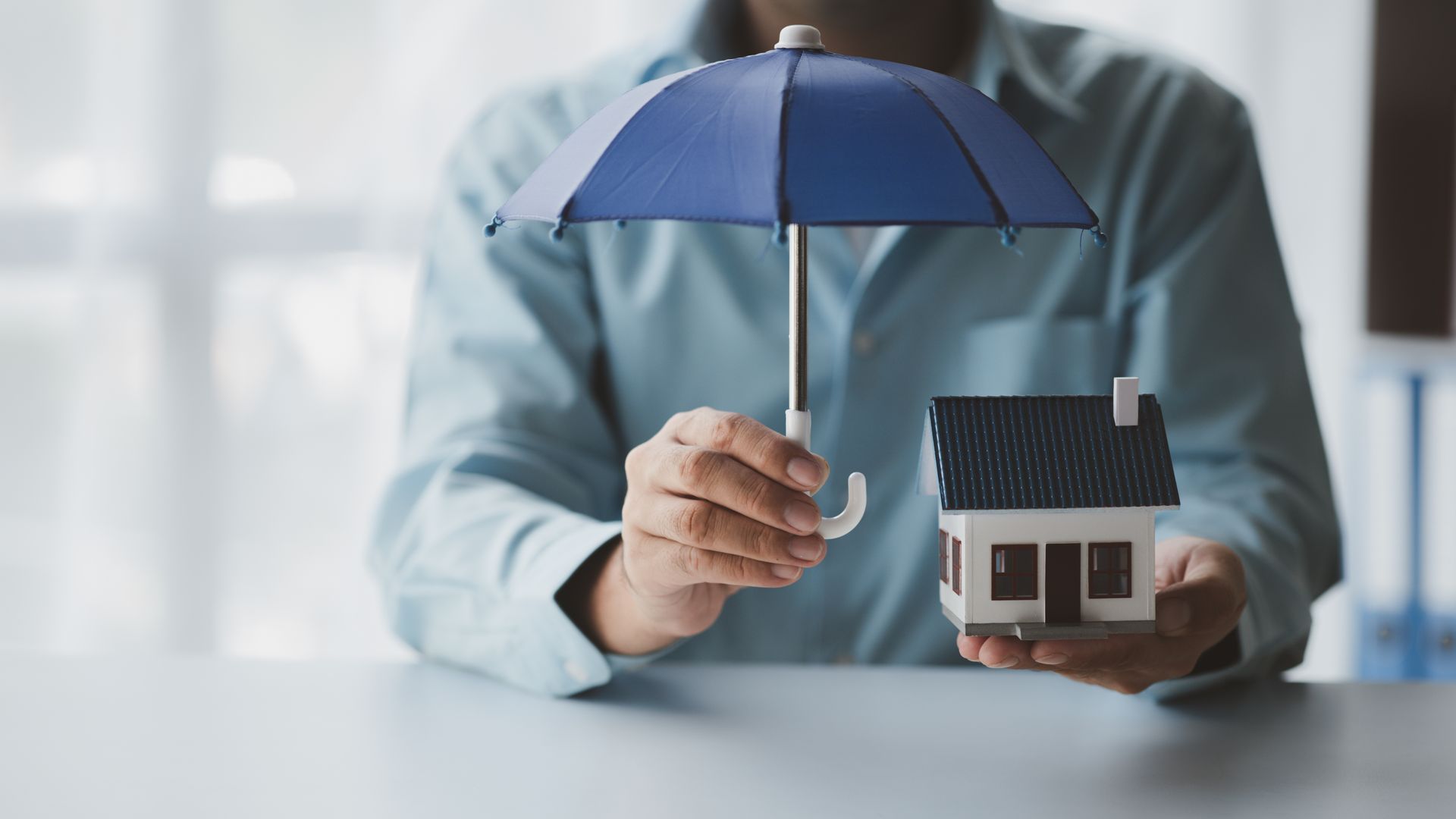 Person holding a blue umbrella over a miniature house, symbolizing home protection.