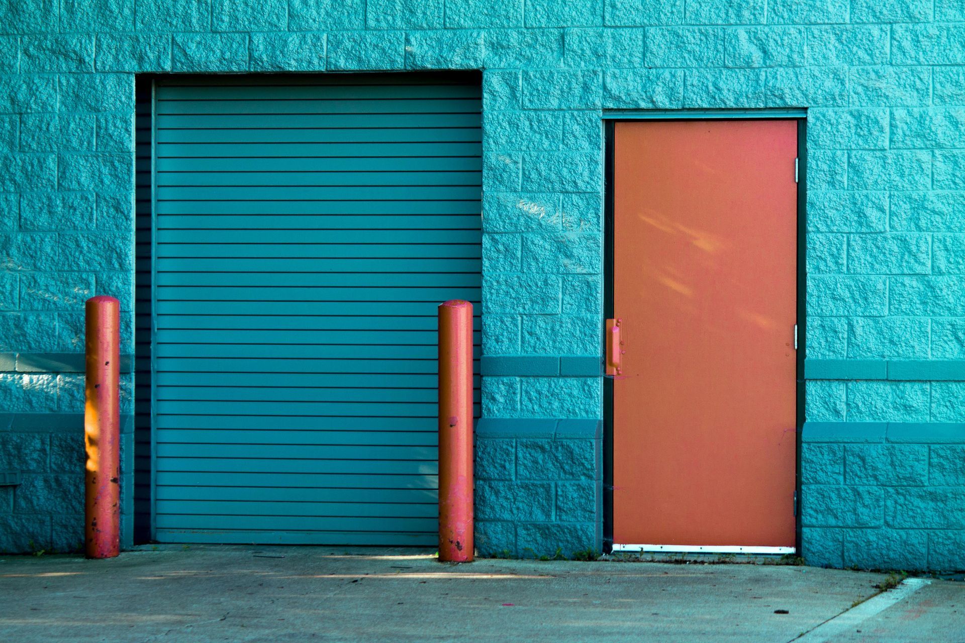 Teal brick building with teal garage door and orange door. Two red posts stand in front.