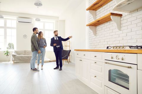 Two people and a realtor in a bright white kitchen, discussing the open shelving and stove Two people and a realtor in a bright white kitchen, discussing the open shelving and stove
