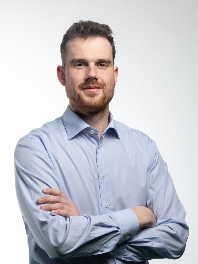 Man with crossed arms, light blue shirt, and a beard, smiling against a white backdrop.