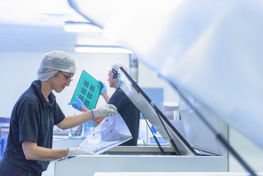 Two people in a clean room setting, wearing hairnets and gloves, handling a transparent sheet and a blue grid.