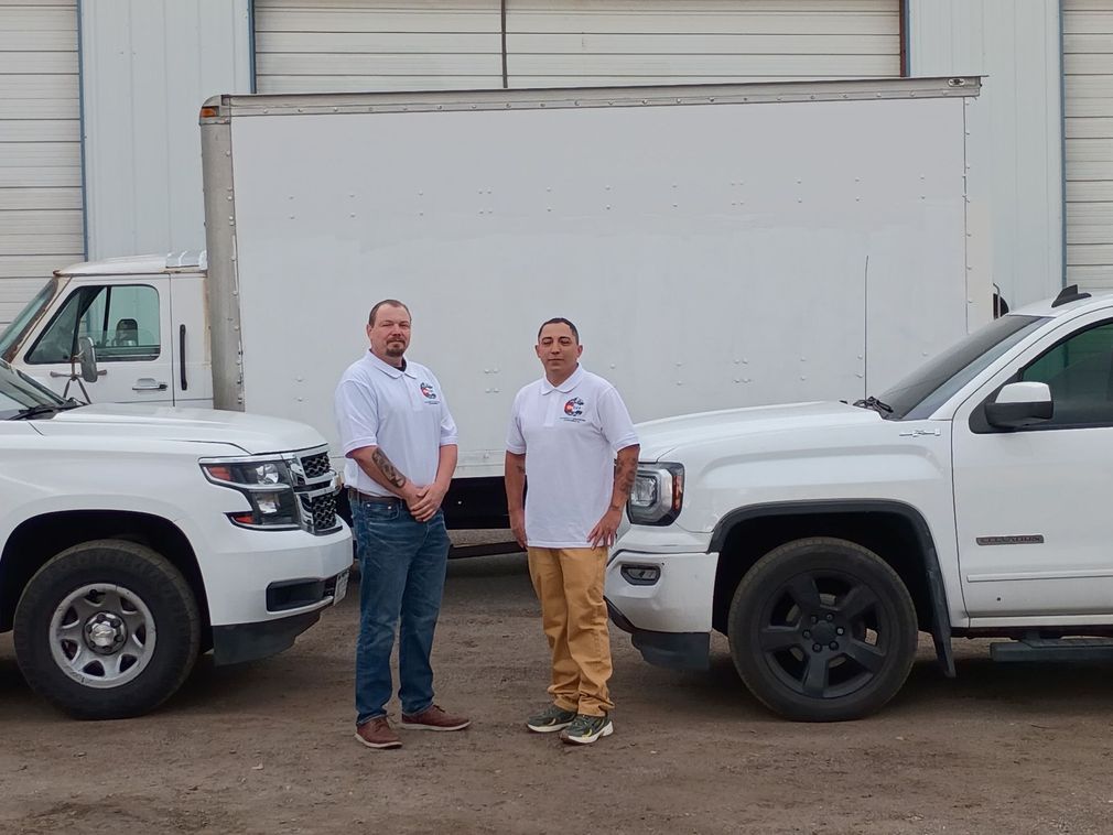 Two men standing between two white pickup trucks in front of a warehouse loading dock
