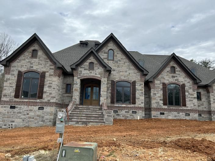 Stone house with gabled roof, front steps, and unfinished yard under a cloudy sky