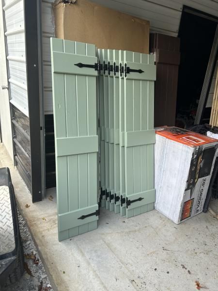 Green shutters leaning against a wall next to a boxed appliance on a concrete porch.