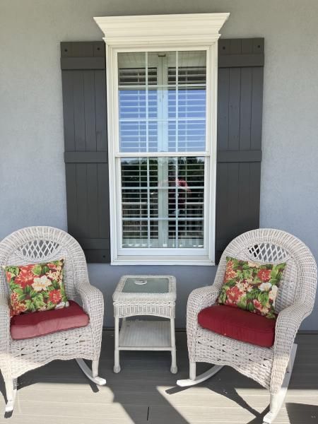 White wicker patio chairs with red cushions flank a small table on a porch with gray shutters and a window.