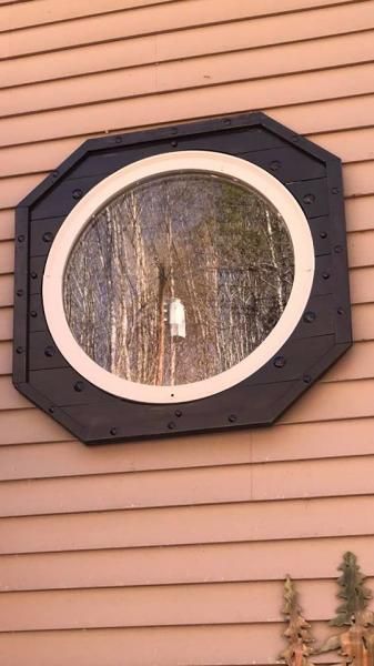 Octagonal black-framed round window on beige siding, reflecting bare trees.
