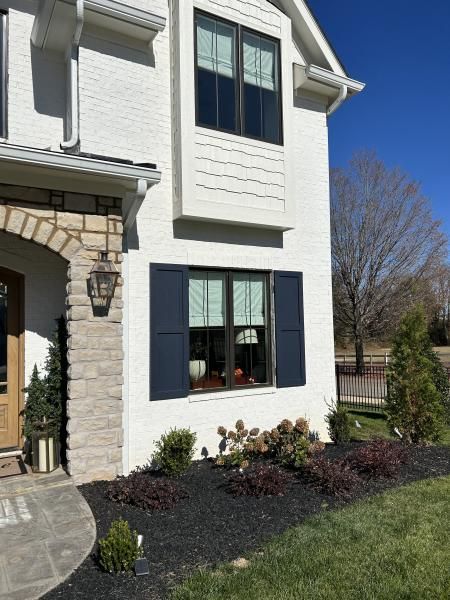 Front of a white house with dark shutters, stone entry, and a small landscaped yard under a clear sky