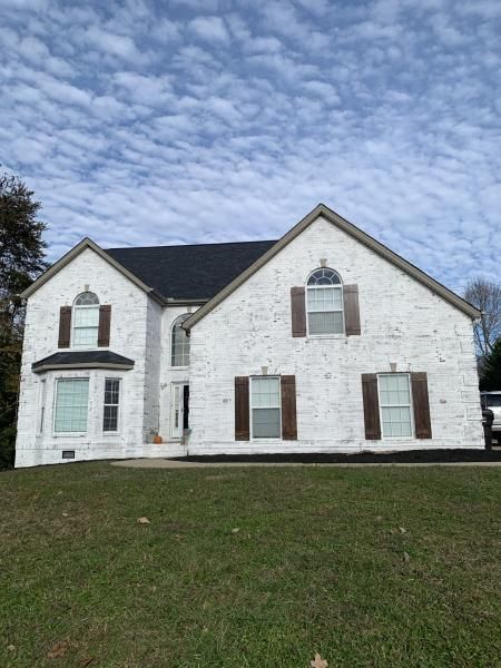 Two-story white stone house with brown shutters and a grassy front lawn under a cloudy sky