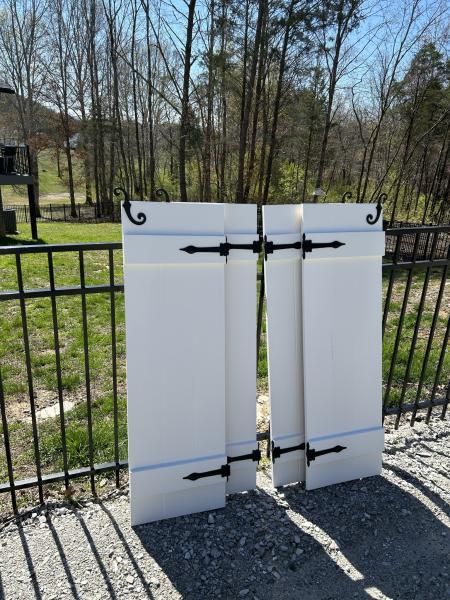 Two white metal gate panels with black hinges, leaning open beside a black fence outdoors.