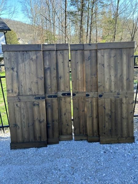 Wooden double gate with black hinges on gravel, set against a fenced yard and leafless trees.