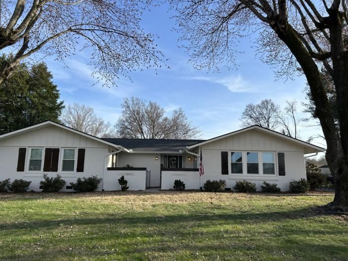 Single-story white house with dark roof and trees in a grassy yard under a blue sky